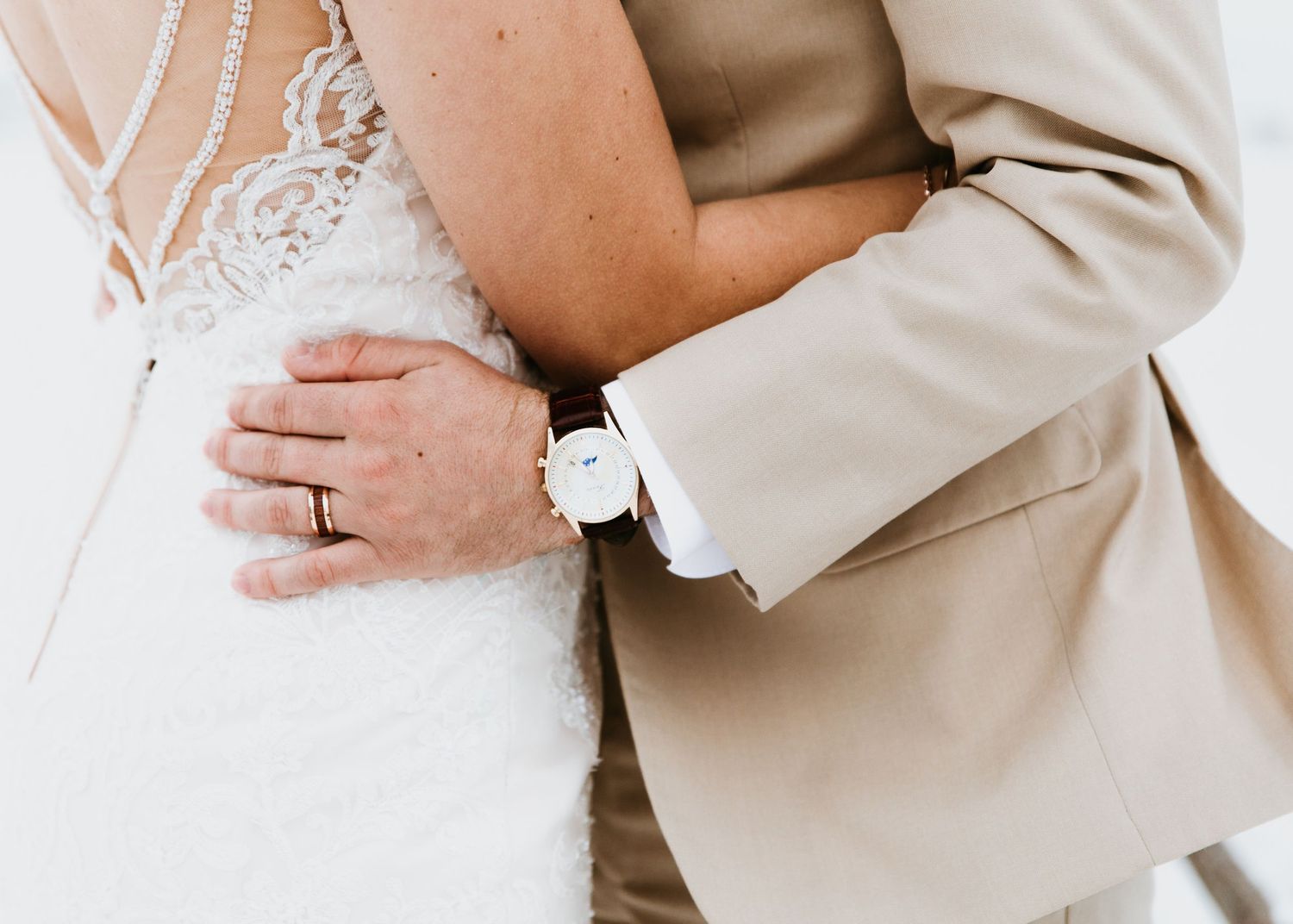 Close-up of groom in tan suit embracing bride in lace dress, groom wearing a white watch.