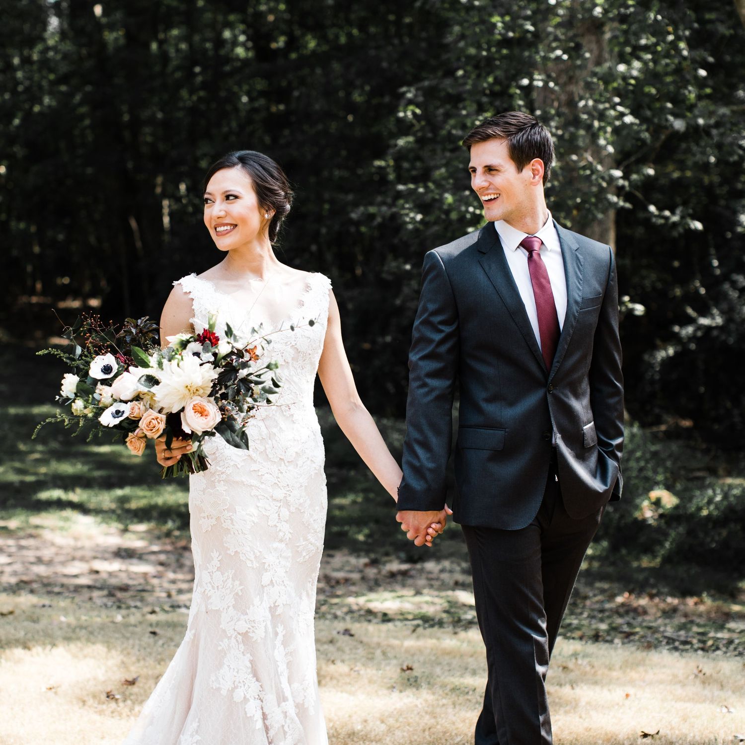 Bride and groom holding hands walking through sunlit wooded path on wedding day.
