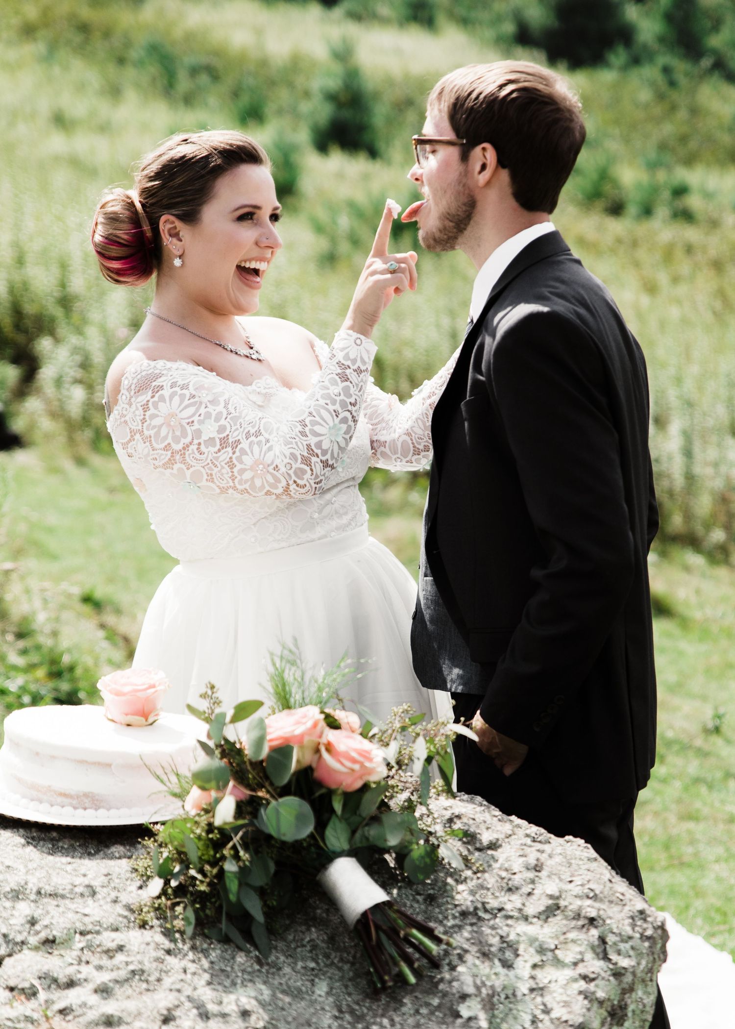 Bride in lace wedding dress feeds cake to groom outdoors with floral bouquet and white cake on stone.