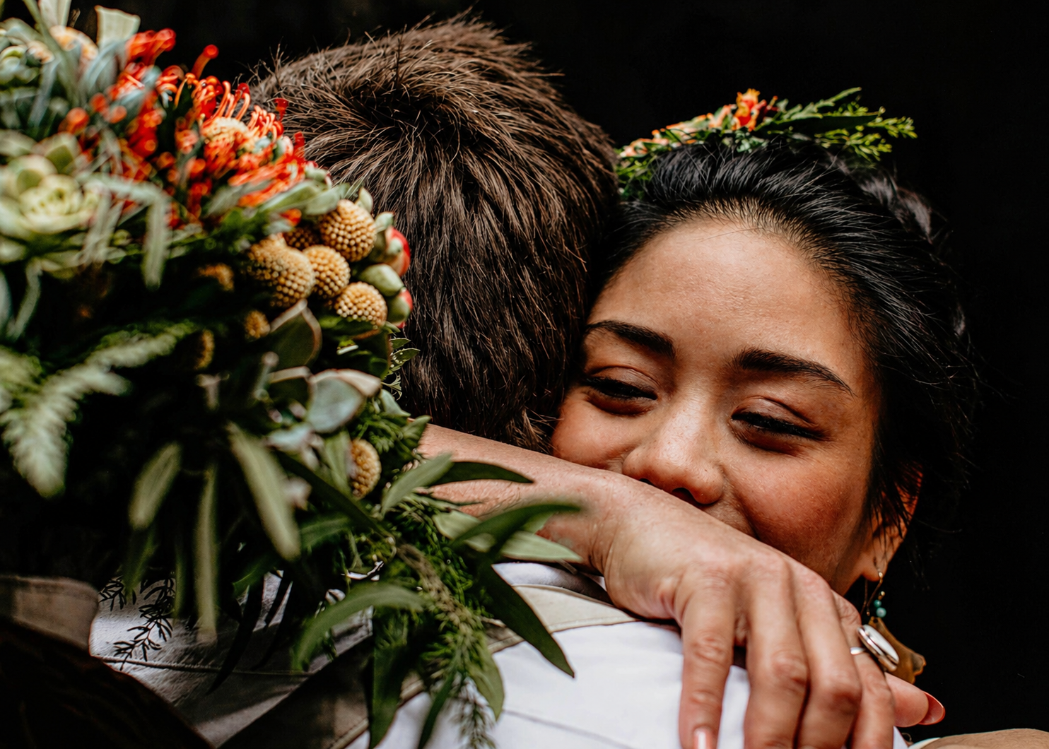 Emotional bride with floral crown hugging someone while holding a vibrant bouquet of tropical flowers.