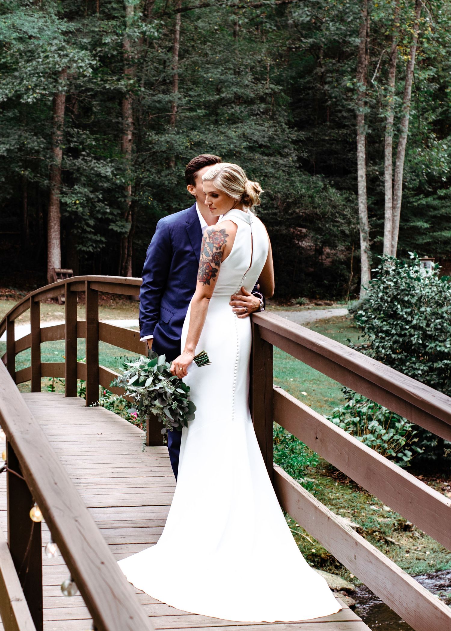 Bride in sleek backless gown and groom embrace on rustic wooden bridge in forest setting.