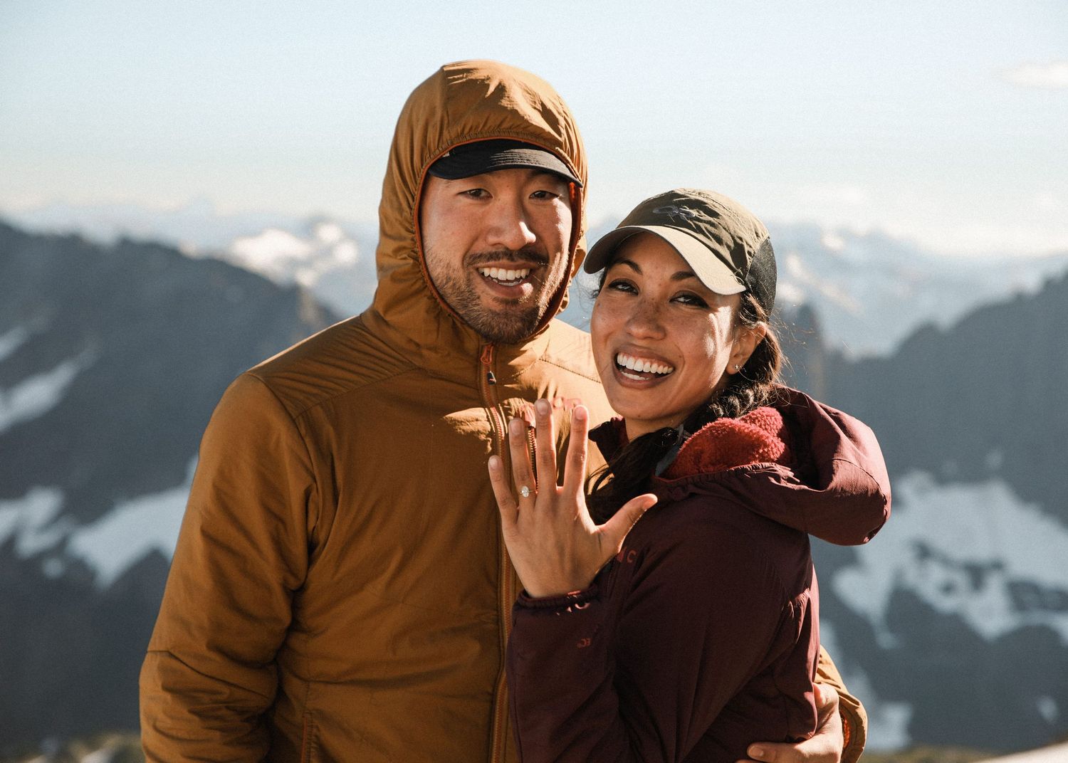 Happy couple showing off engagement ring on snowy mountain summit wearing winter jackets.