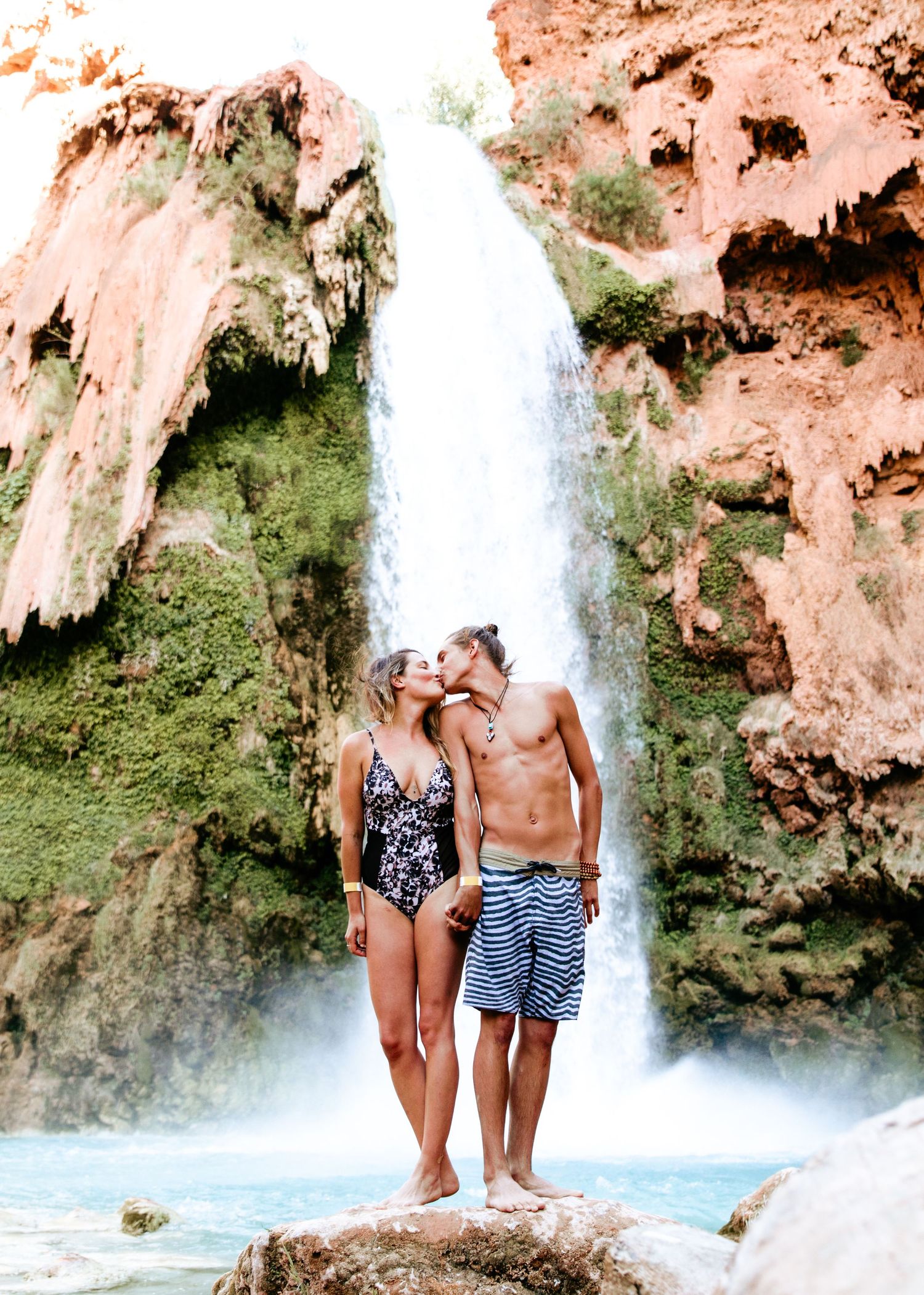 Couple kissing in swimwear on rocks in front of a stunning waterfall.