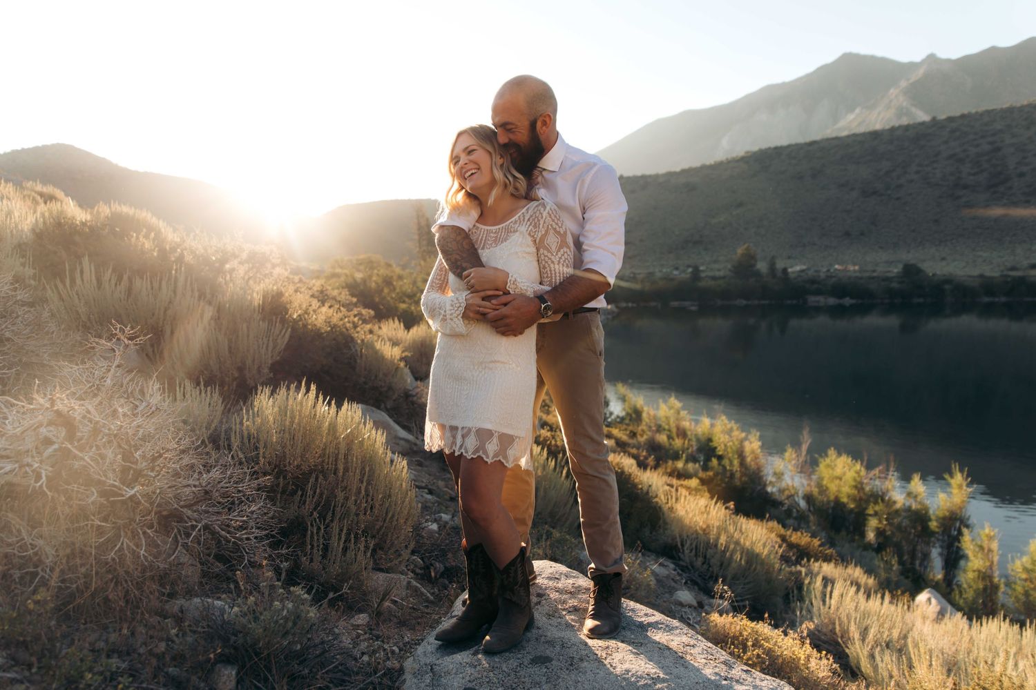 Couple embracing at sunset by a mountain lake, woman in white lace dress.