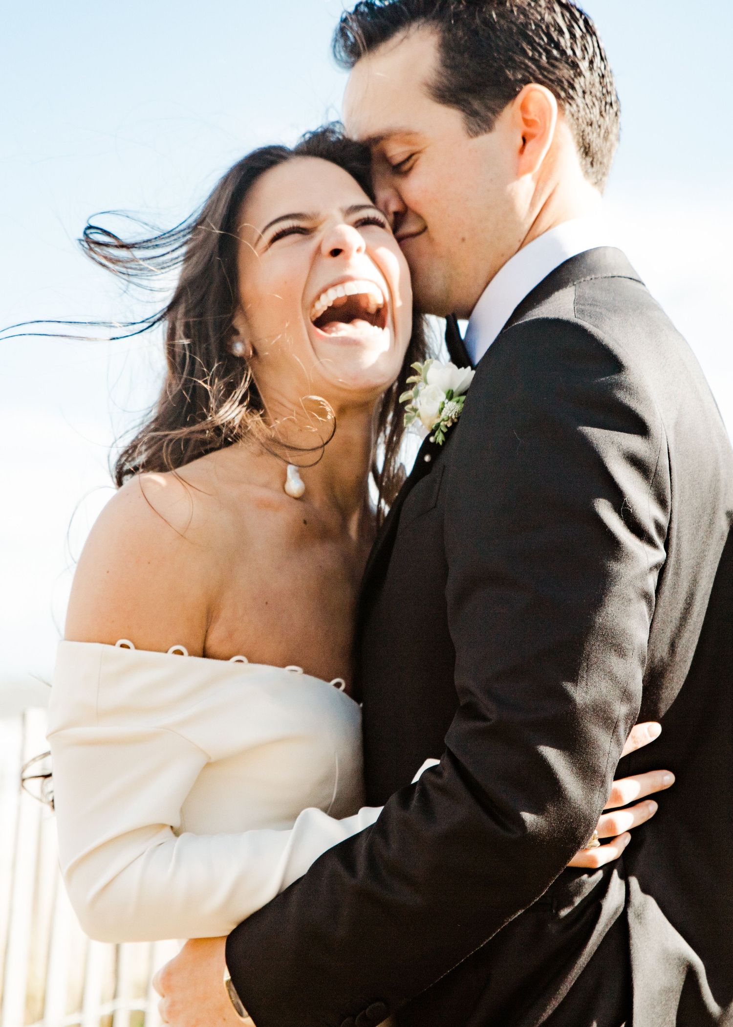 Joyful bride in white off-shoulder gown laughs as groom in black suit kisses her cheek outdoors.
