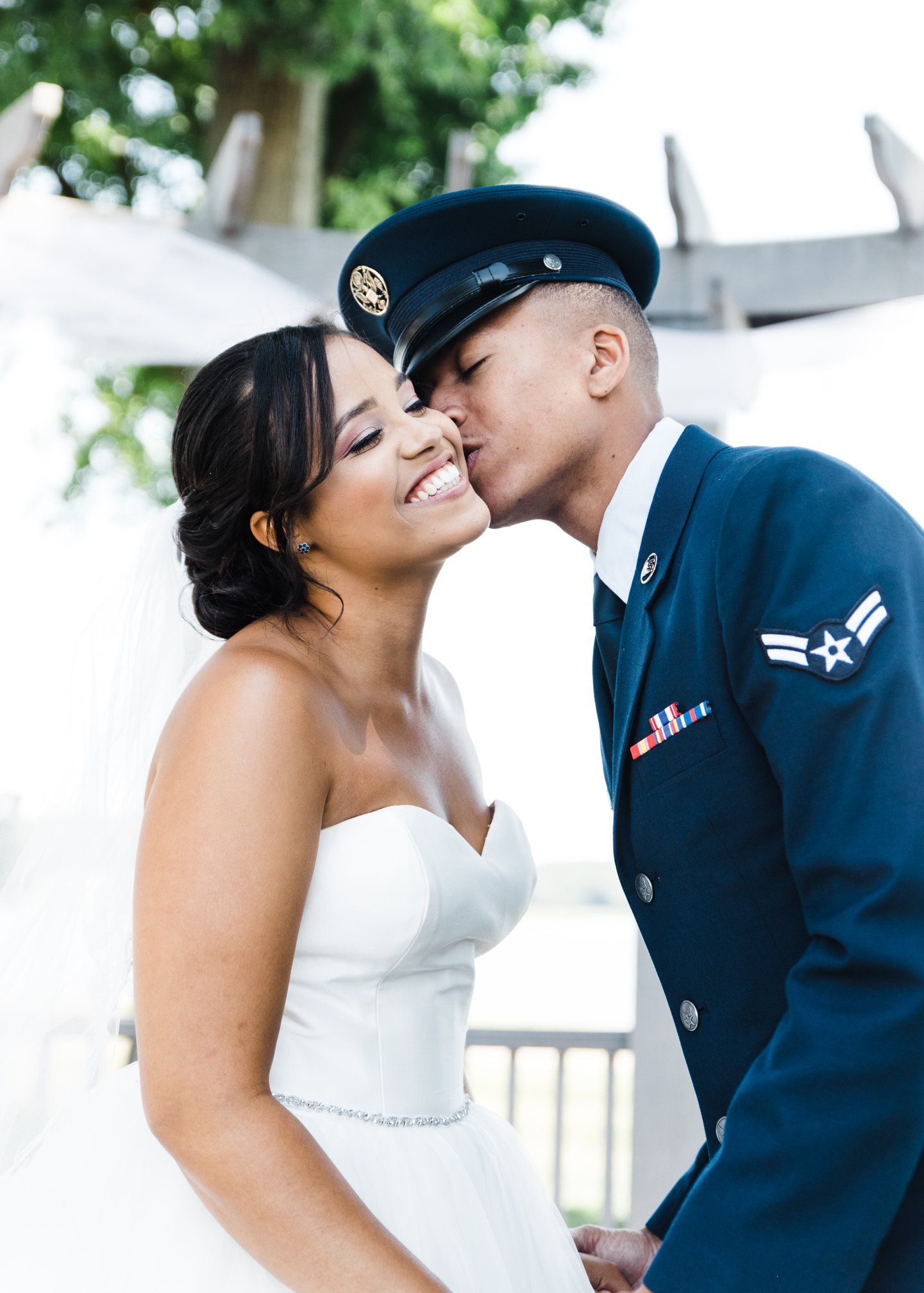 Military groom in Air Force dress uniform kisses smiling bride on cheek outdoors.