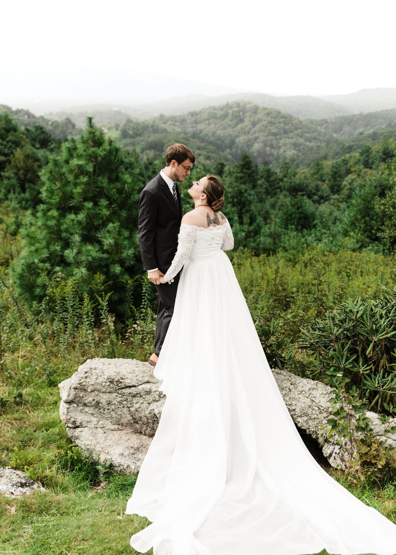 Bride and groom share a tender moment on a rocky overlook surrounded by lush green mountains.