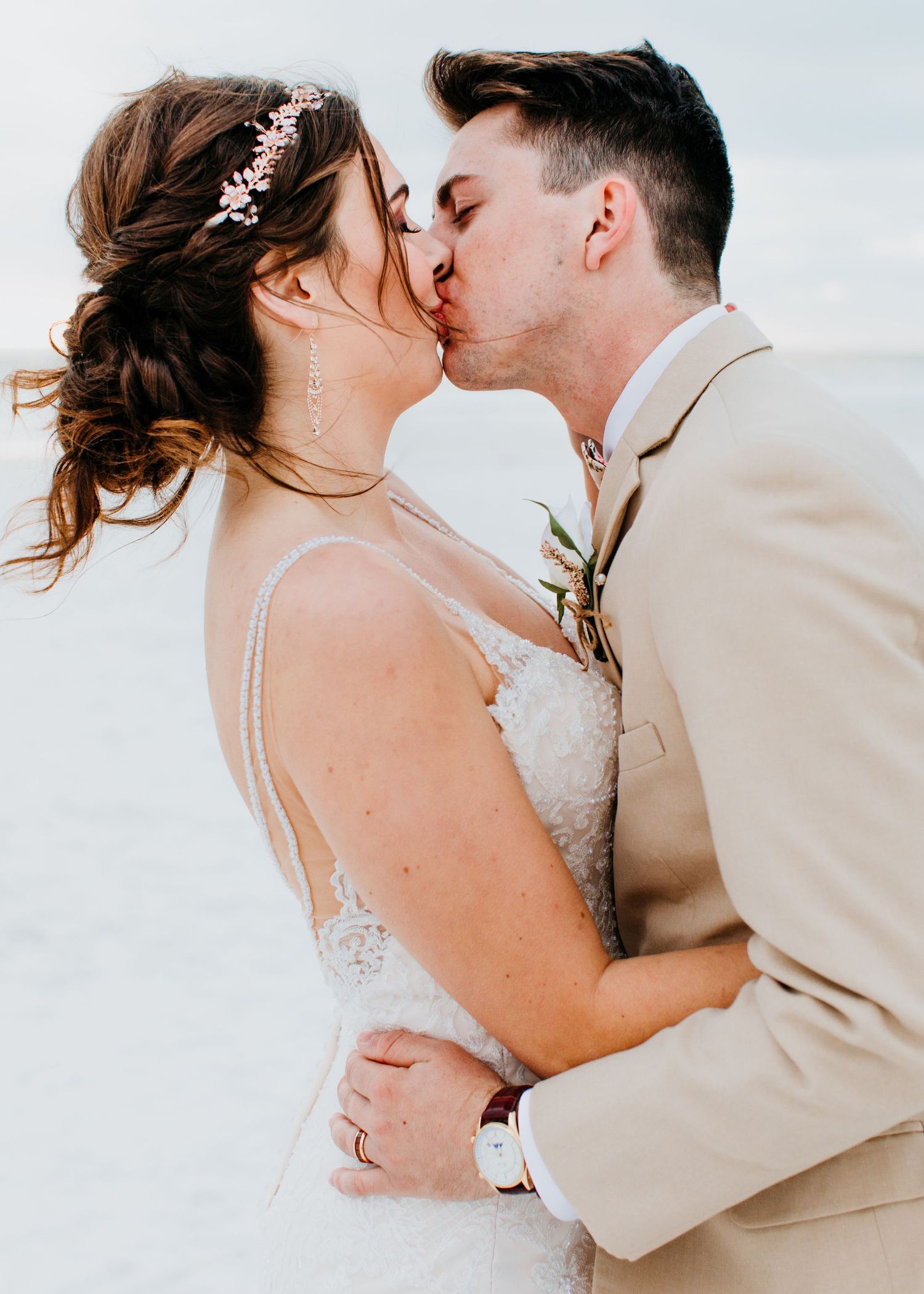 Bride and groom sharing passionate kiss on beach with soft ocean background at sunset.