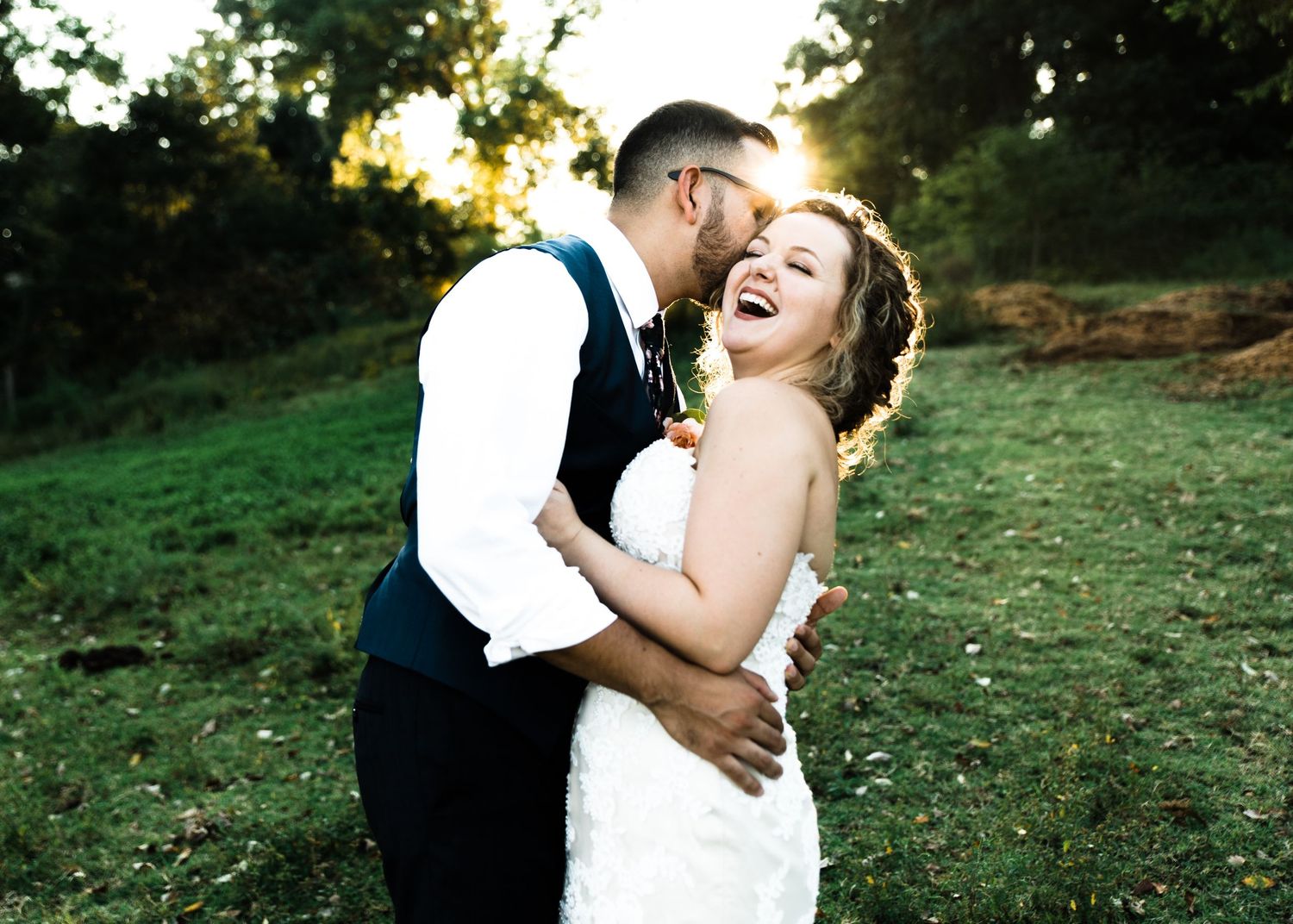 Groom kissing laughing bride on cheek in sunny outdoor wedding photo.