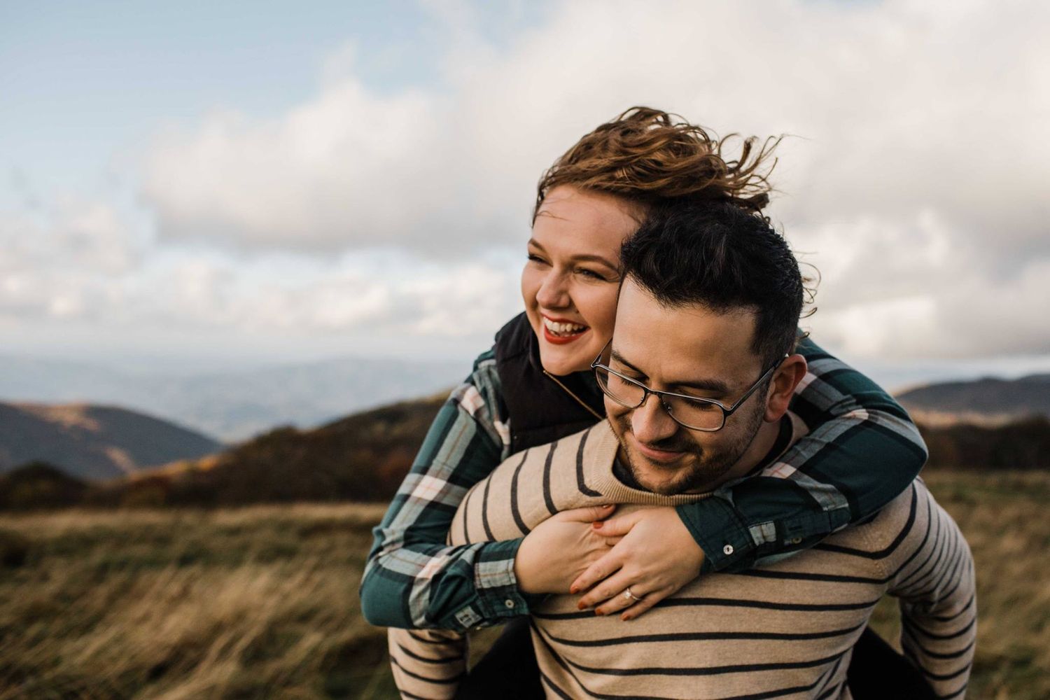 Woman hugging man from behind as he gives her a piggyback ride on a breezy mountain hillside.