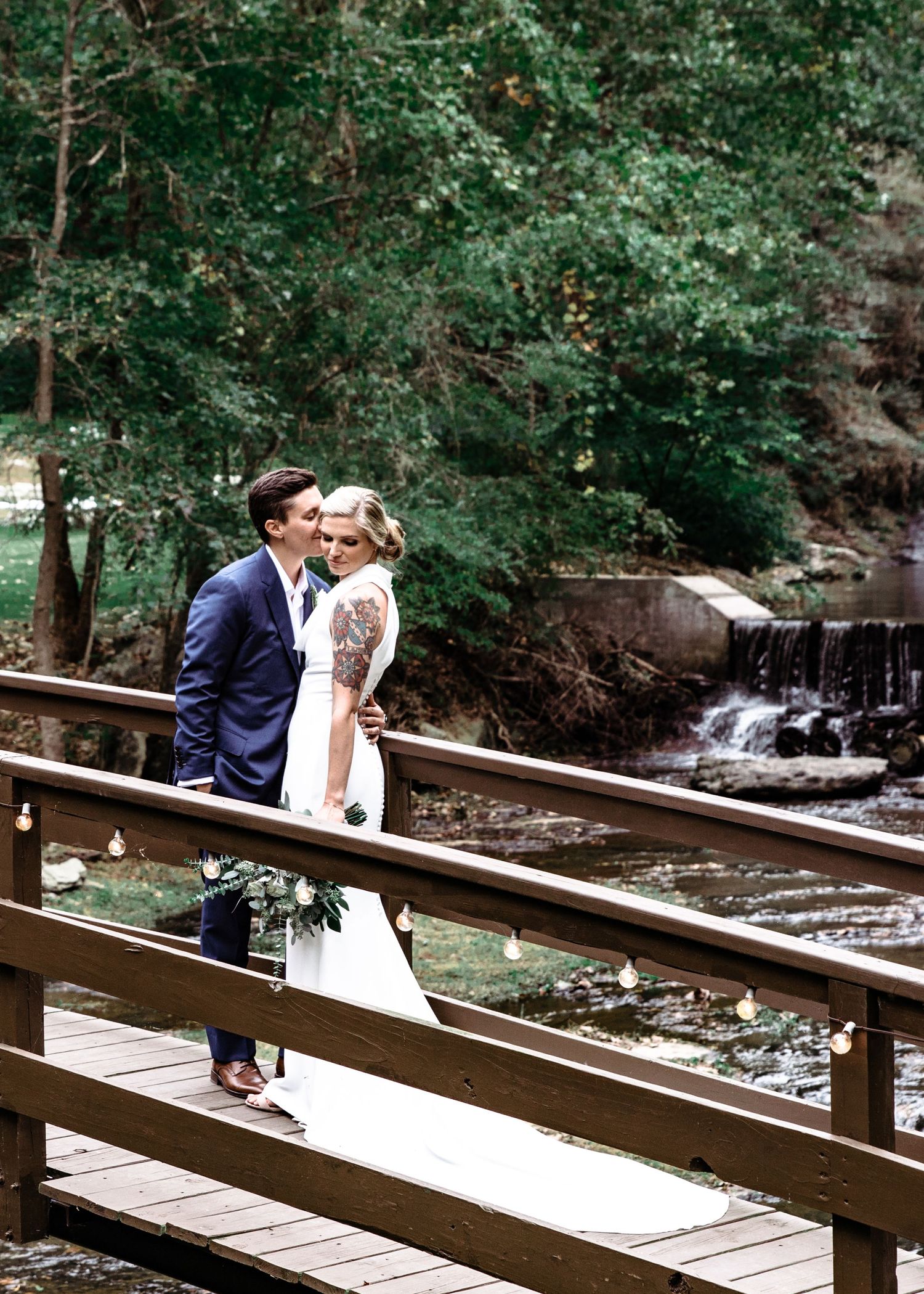 Bride and groom sharing romantic moment on wooden bridge with scenic waterfall backdrop.