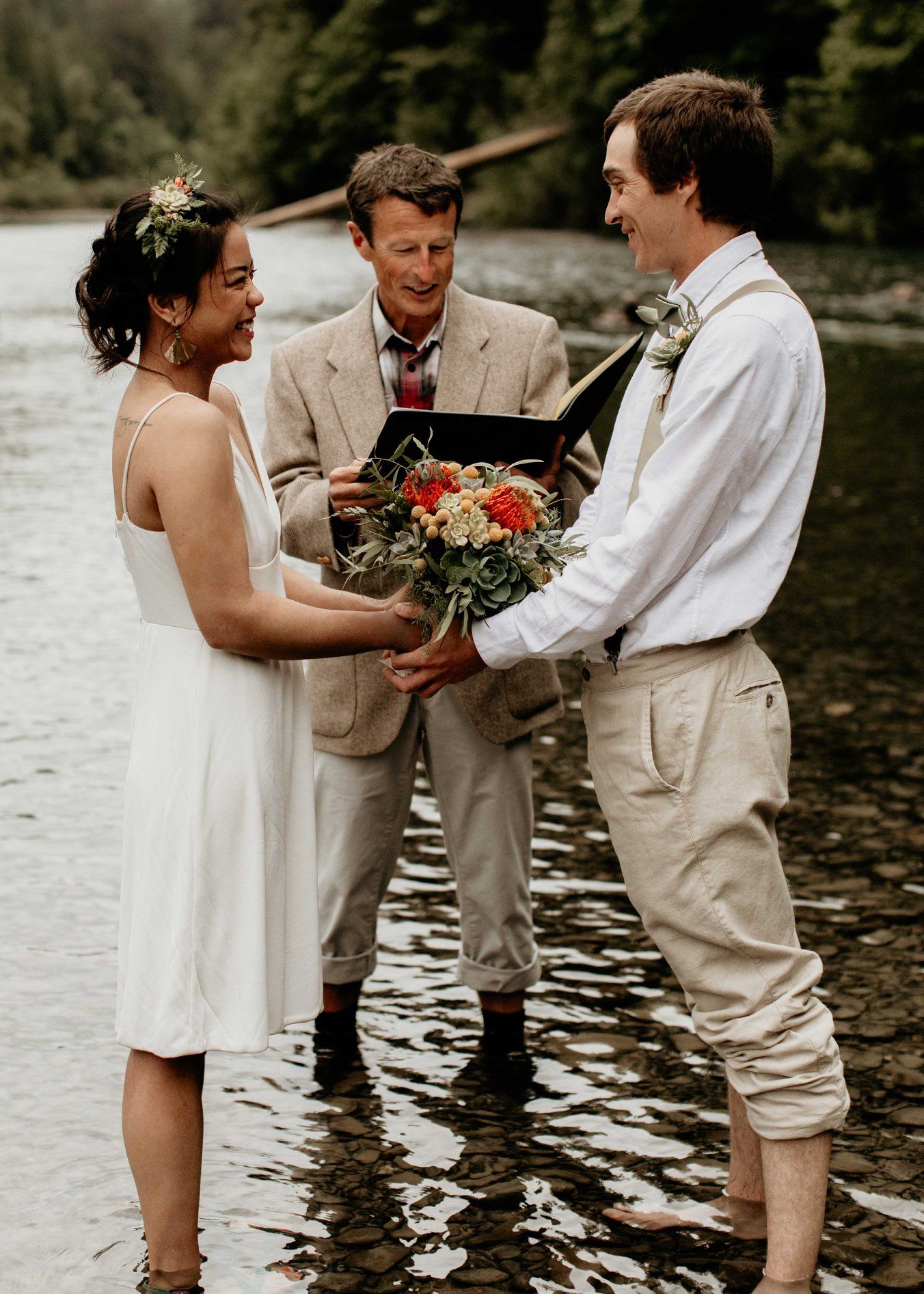 Outdoor riverside wedding ceremony with bride in white dress, groom in linen, and officiant holding a book.