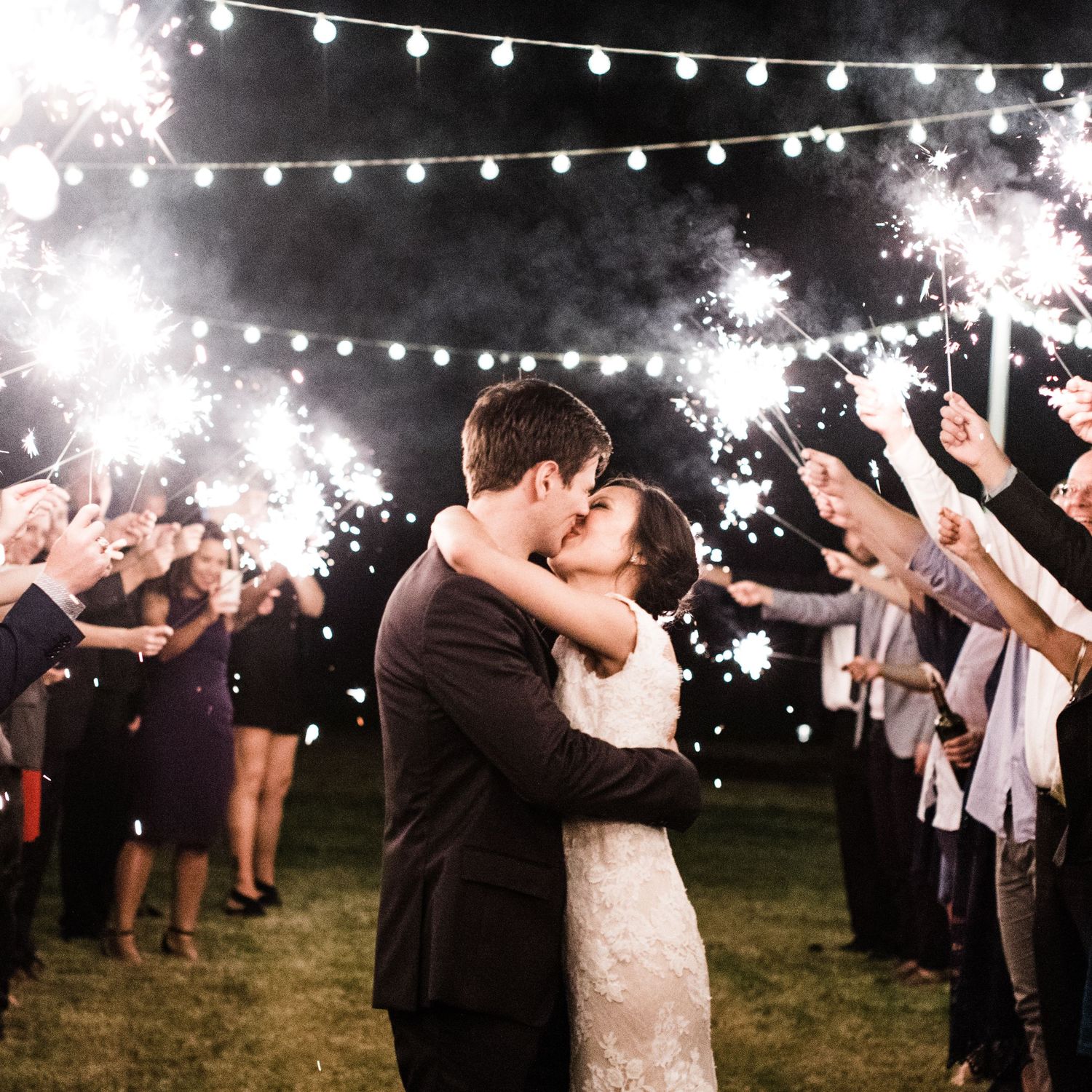 Bride and groom sharing a first kiss during a magical sparkler exit surrounded by cheering wedding guests.