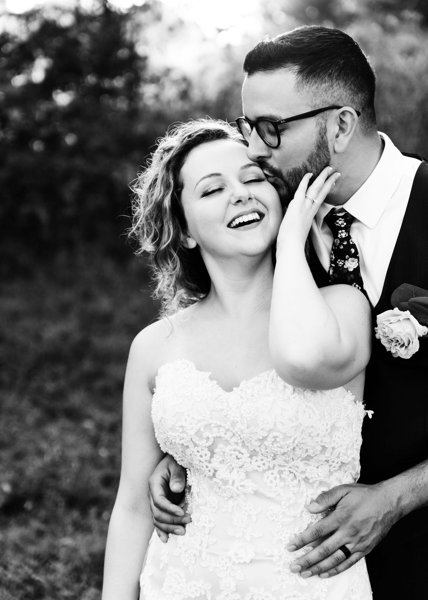 Black and white photo of smiling bride in lace dress as groom tenderly kisses her cheek.