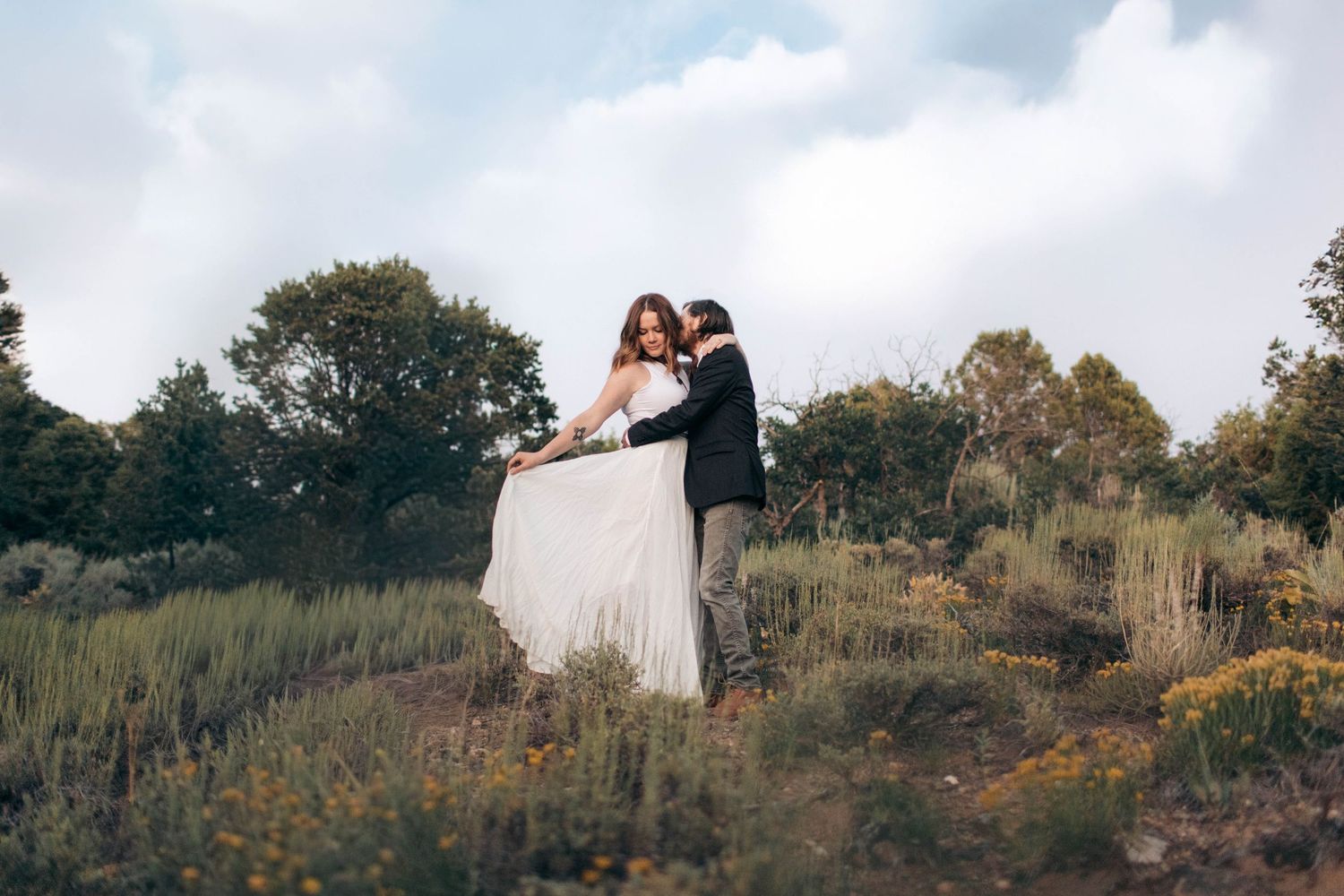 Elegant couple in wedding attire dance together in a lush wildflower garden under a cloudy sky.