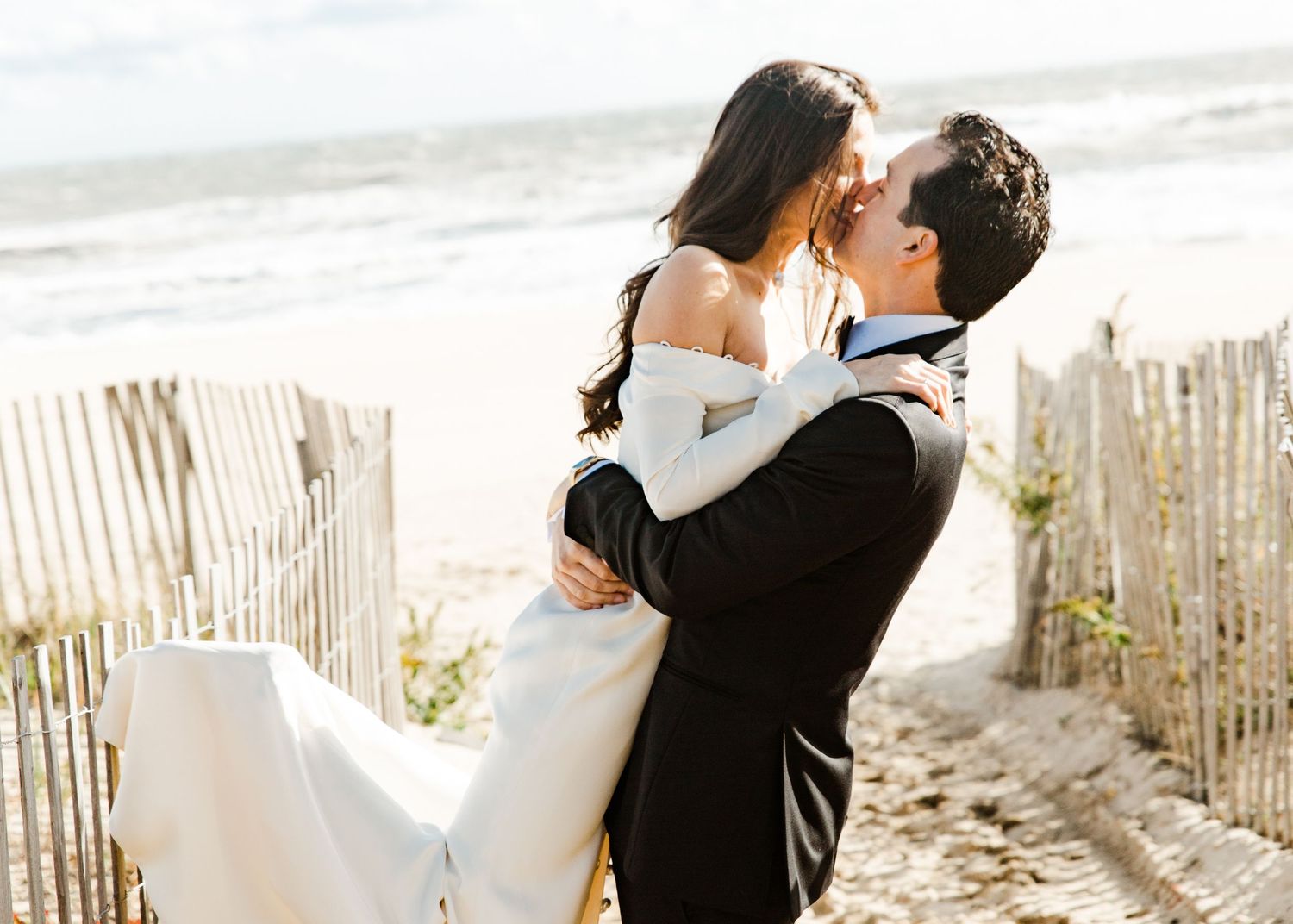Groom lifting bride for a kiss between sandy beach fence posts with ocean waves in the background.