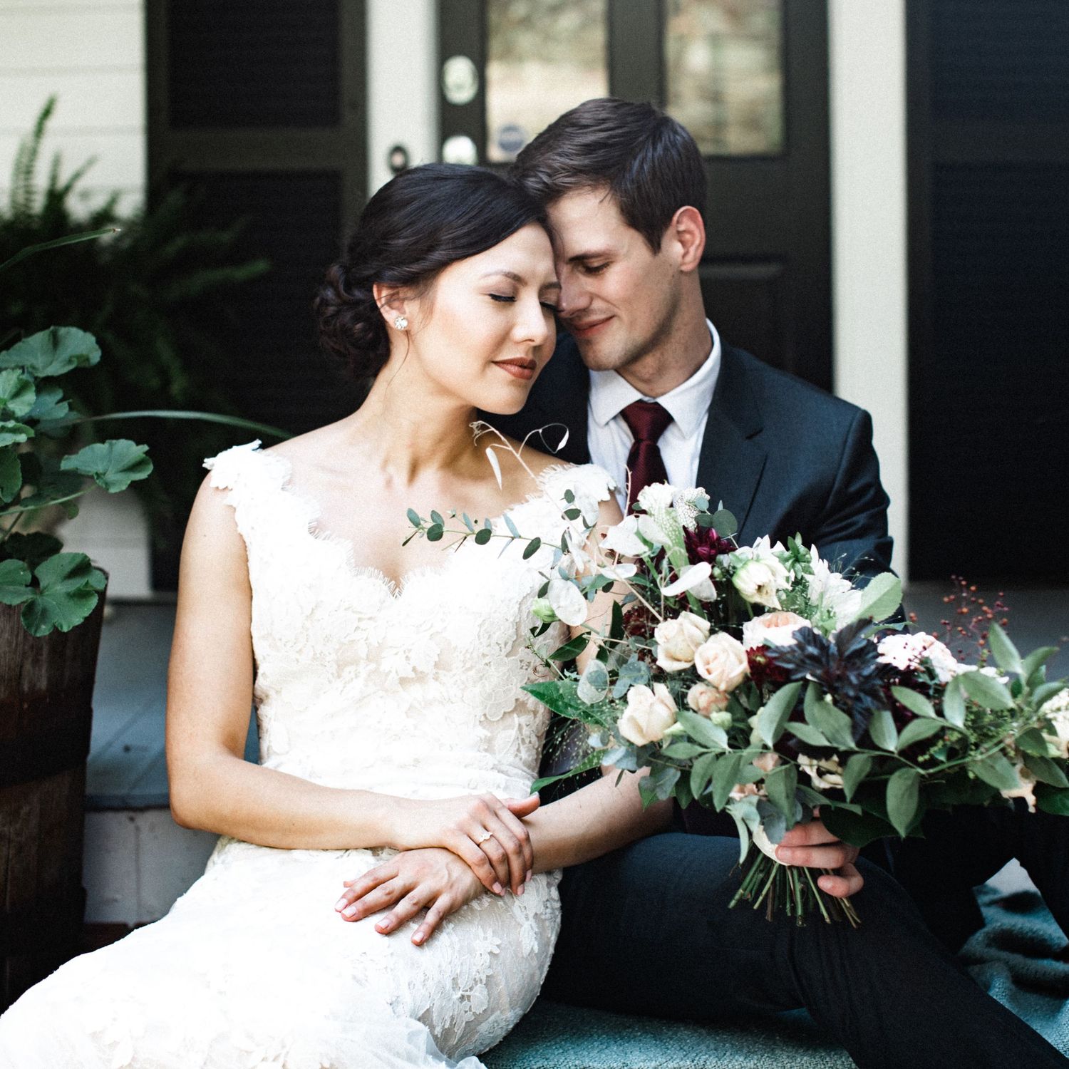 Bride in lace gown holding bouquet with groom in dark suit on porch steps.
