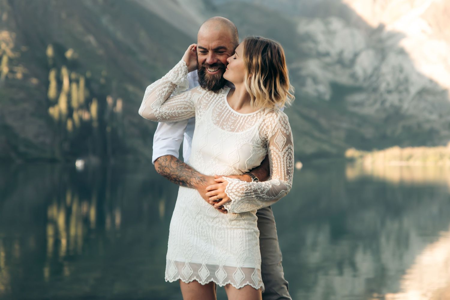 Couple embracing and smiling by a serene alpine lake with mountain reflections in background.