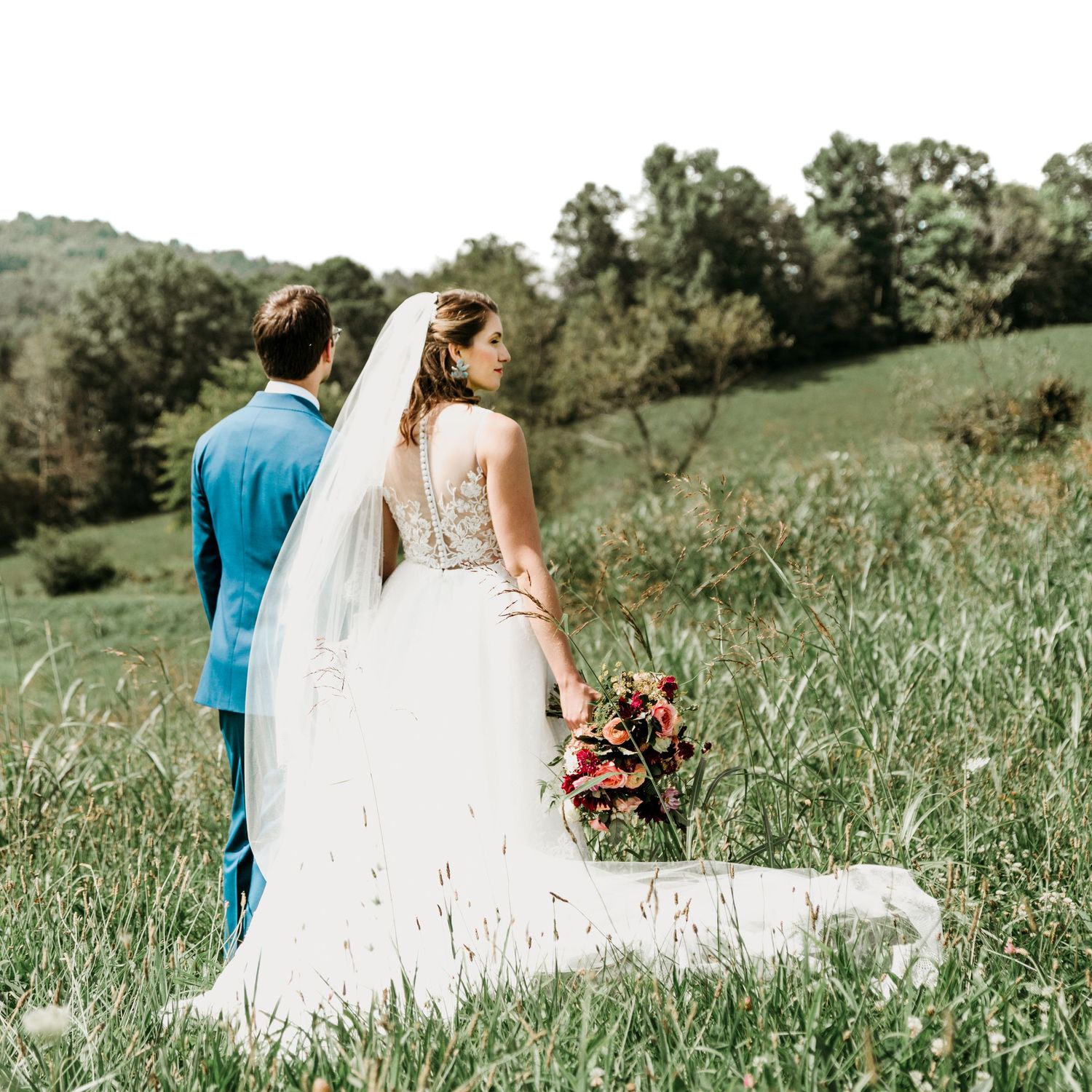 Bride and groom walk hand in hand through tall grass with rolling green hills in the background.
