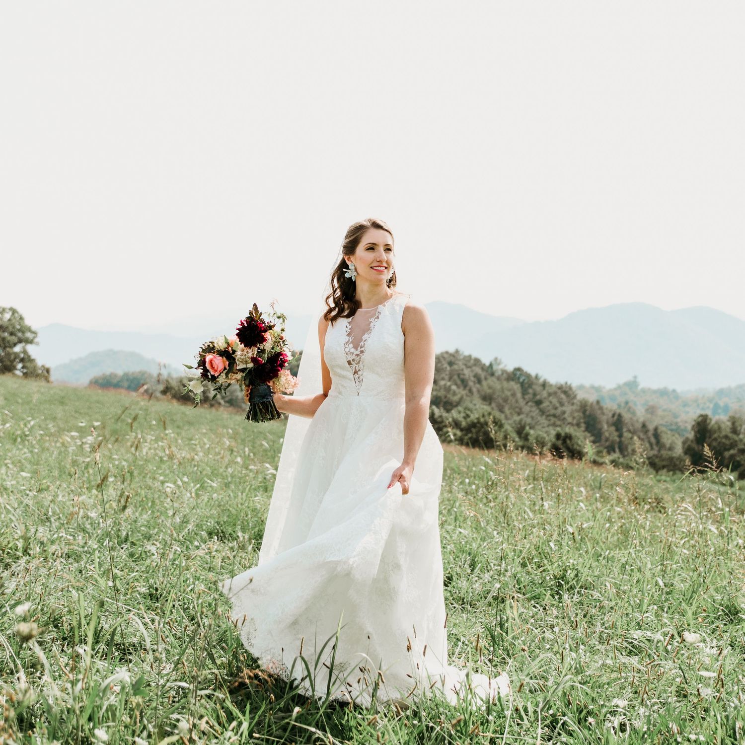 Elegant bride in a flowing white gown holding a burgundy bouquet while walking through a scenic mountain meadow.