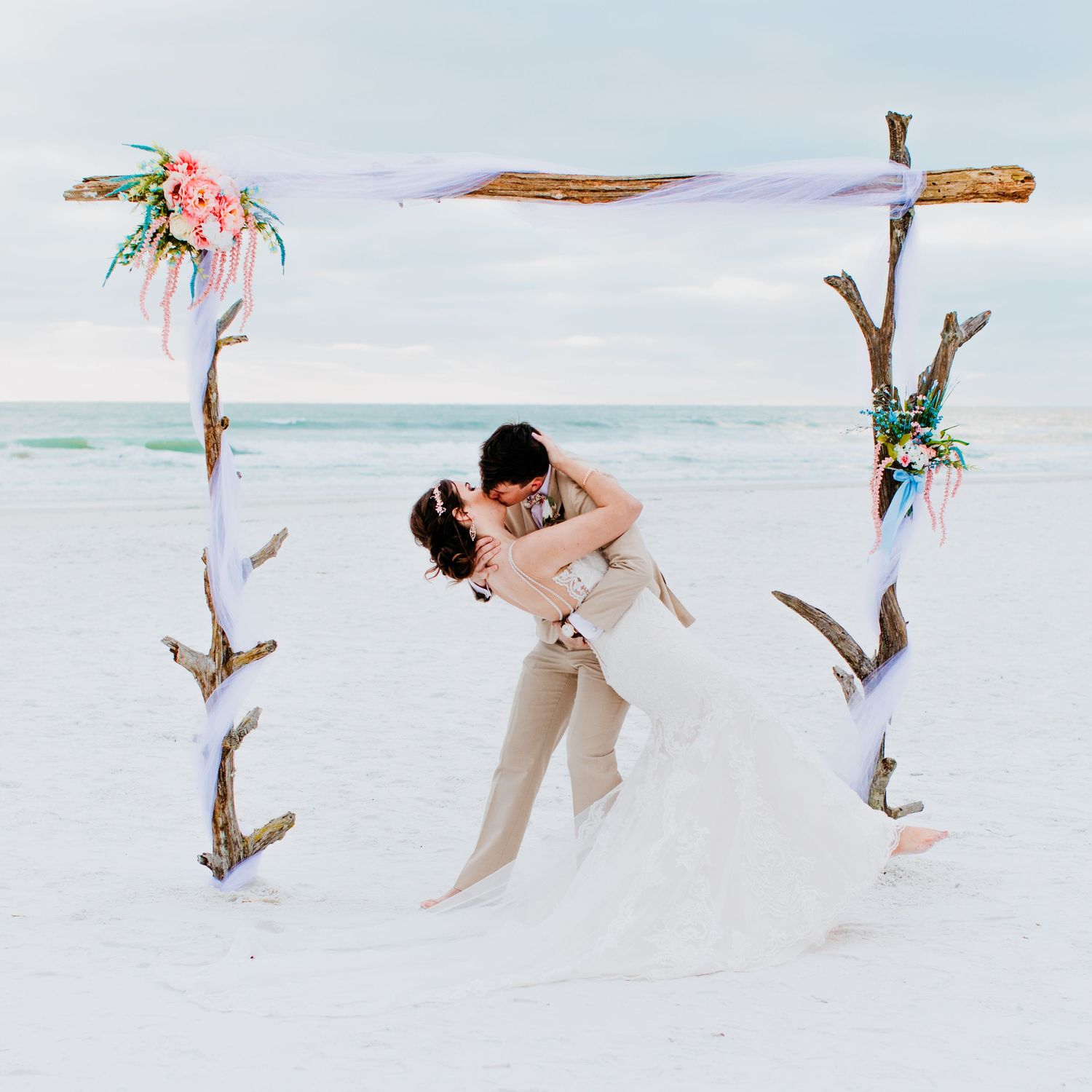 Bride and groom sharing romantic dip kiss under rustic driftwood arch on white sand beach.