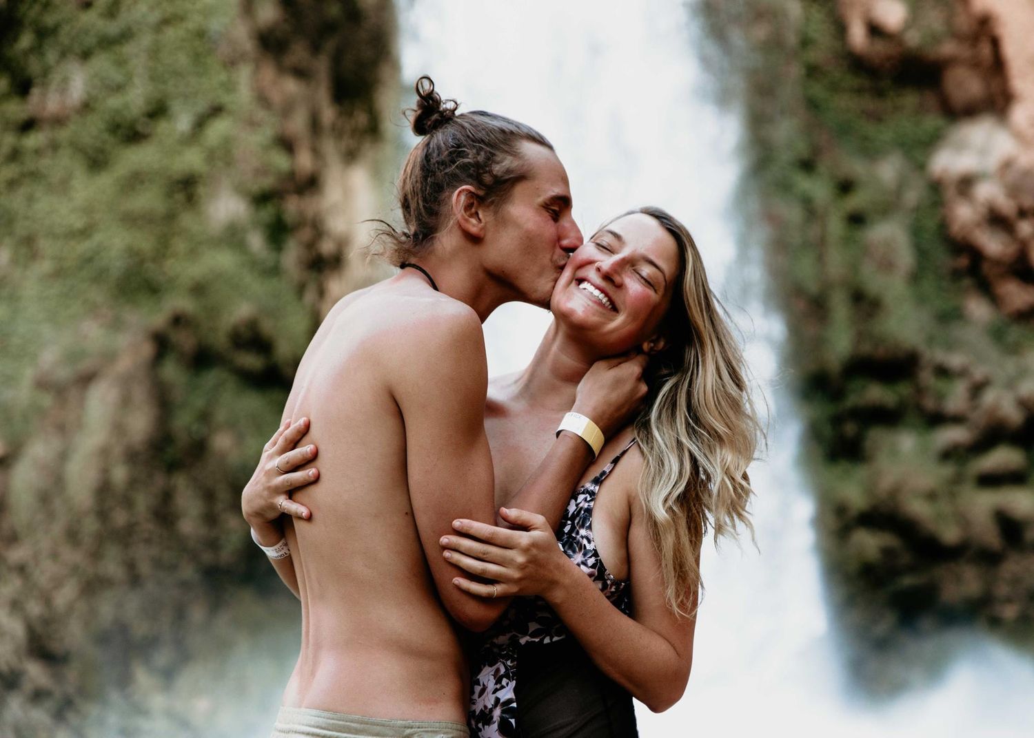 Couple sharing a romantic kiss near a waterfall, man shirtless and woman in floral swimsuit embracing joyfully.