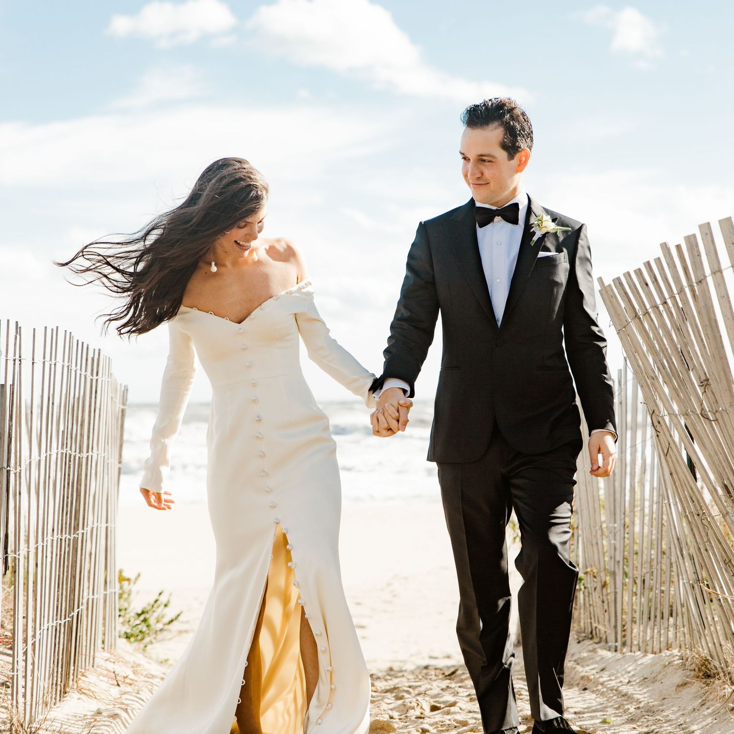 Bride in off-shoulder white gown and groom in black tuxedo hold hands walking on a beach path.