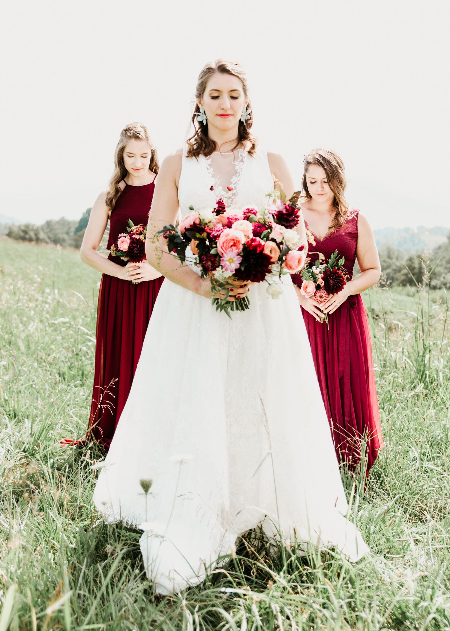 Bride in white gown holds colorful bouquet flanked by two bridesmaids in burgundy dresses in a green field.