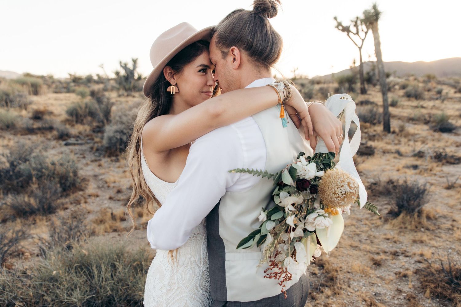 Two brides embracing in Joshua Tree desert landscape, one holding a boho wildflower bouquet.