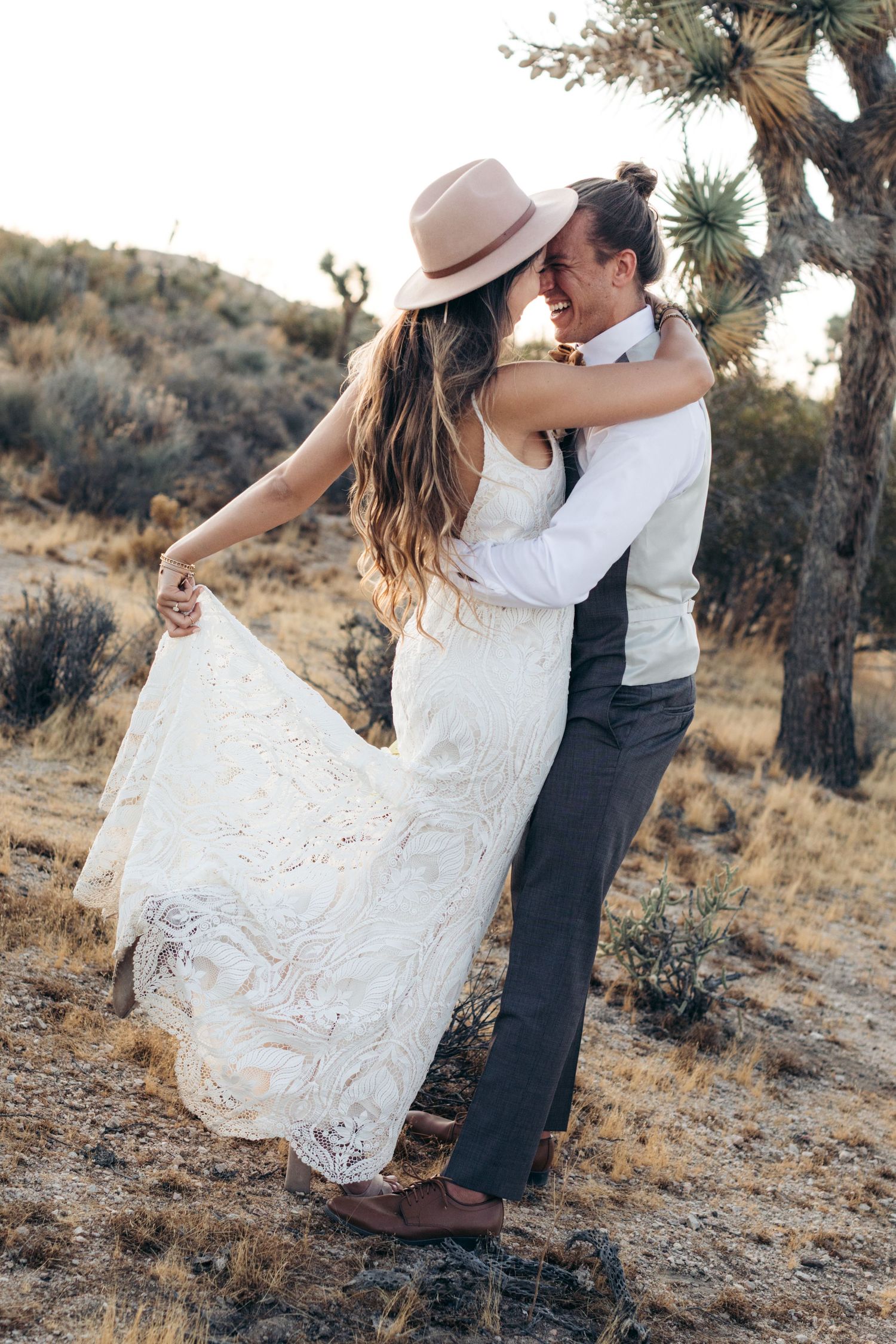 Bride in lace dress and hat embracing groom among desert Joshua trees.