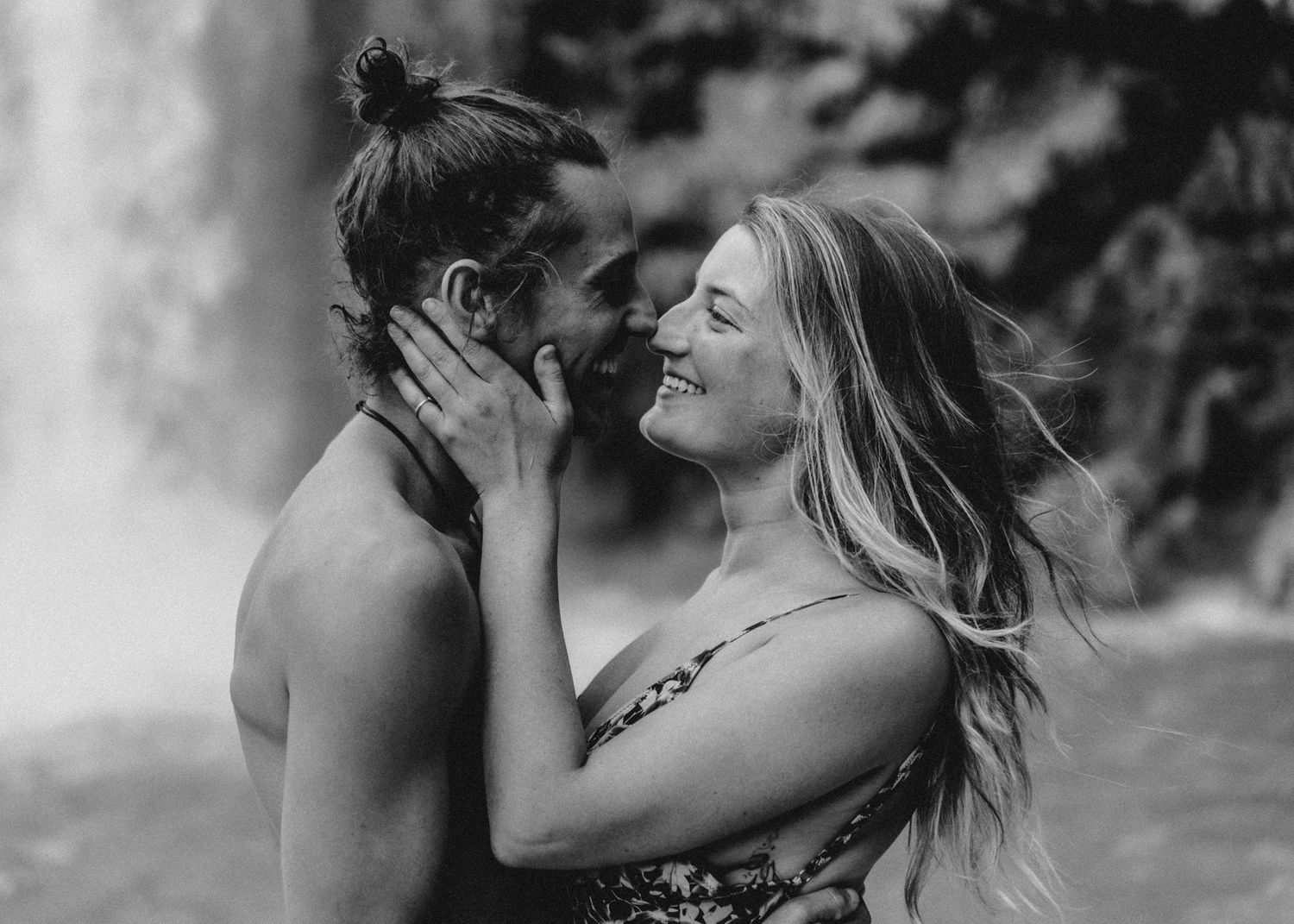 Black and white photo of romantic couple nearly kissing near a waterfall, woman smiling with flowing hair.
