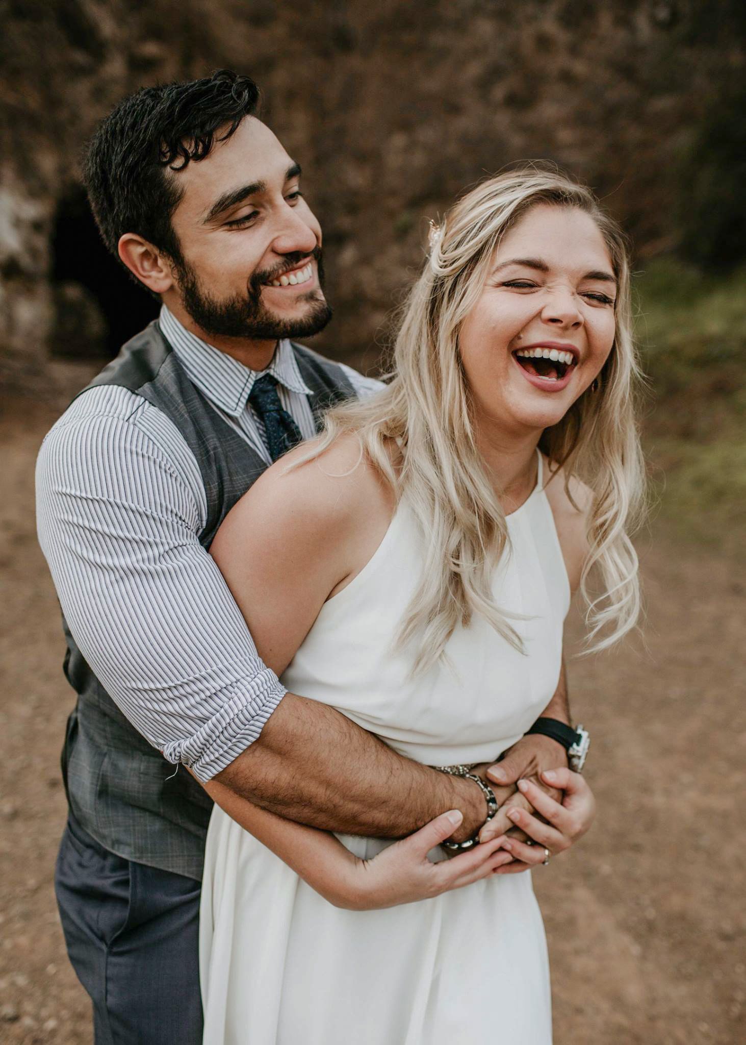 Happy couple embracing outdoors, groom in vest hugging laughing bride in white dress during photo shoot.