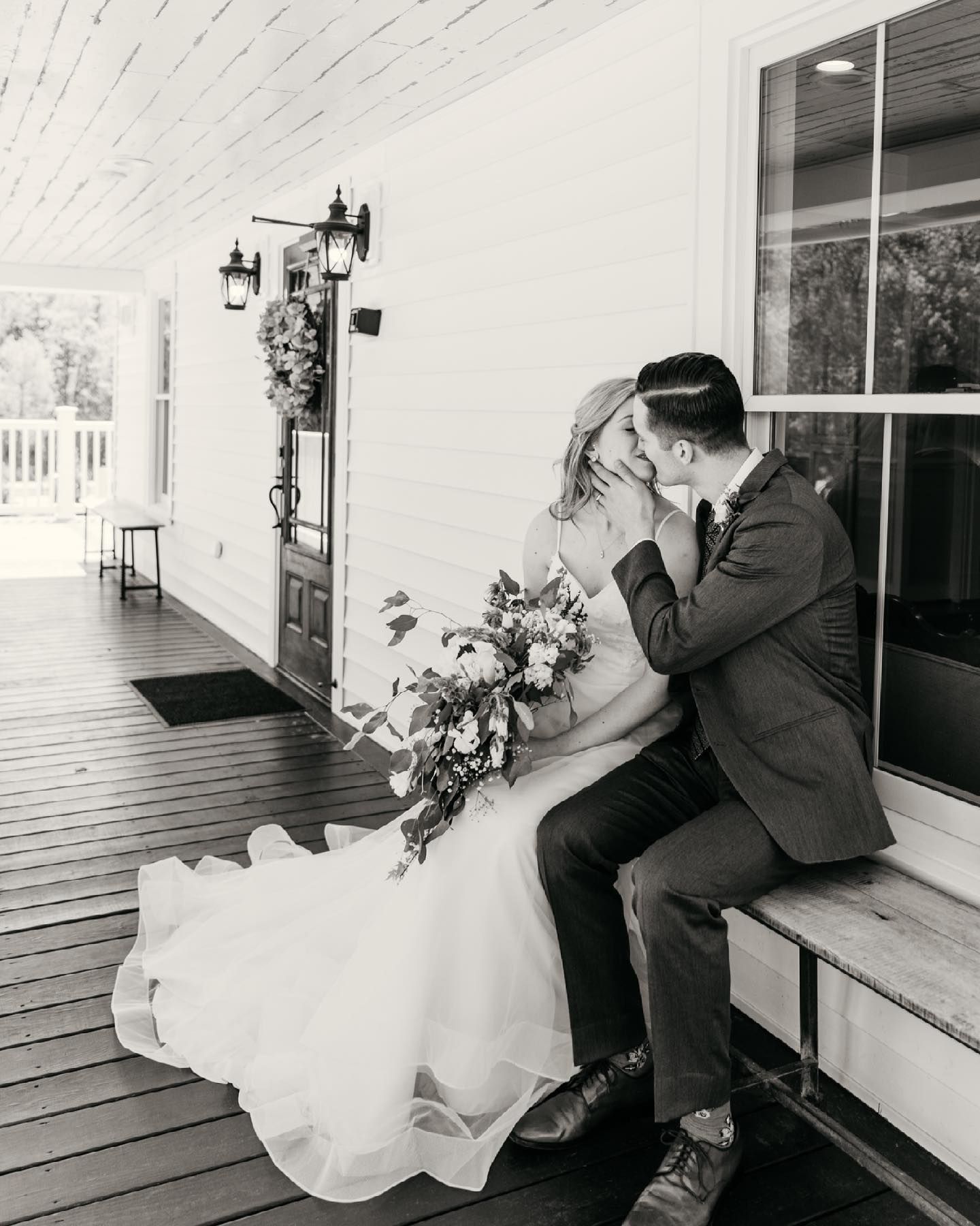 Black and white photo of bride and groom kissing on rustic porch with bouquet.