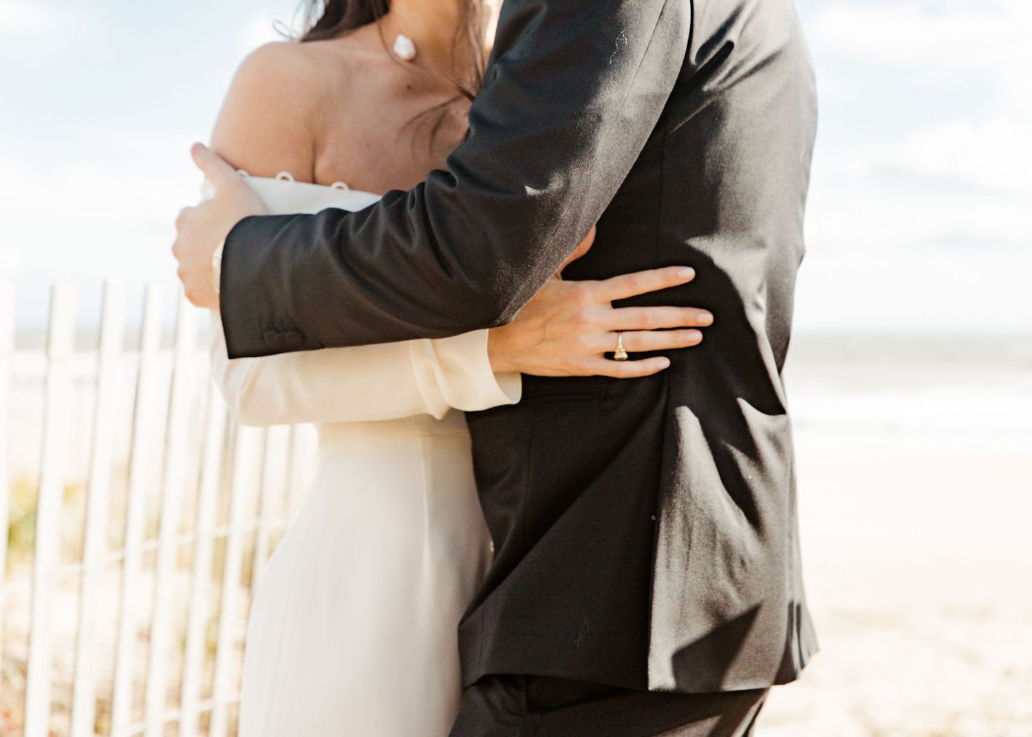 Close-up of bride and groom embracing on the beach, showcasing her diamond engagement ring and wedding dress.