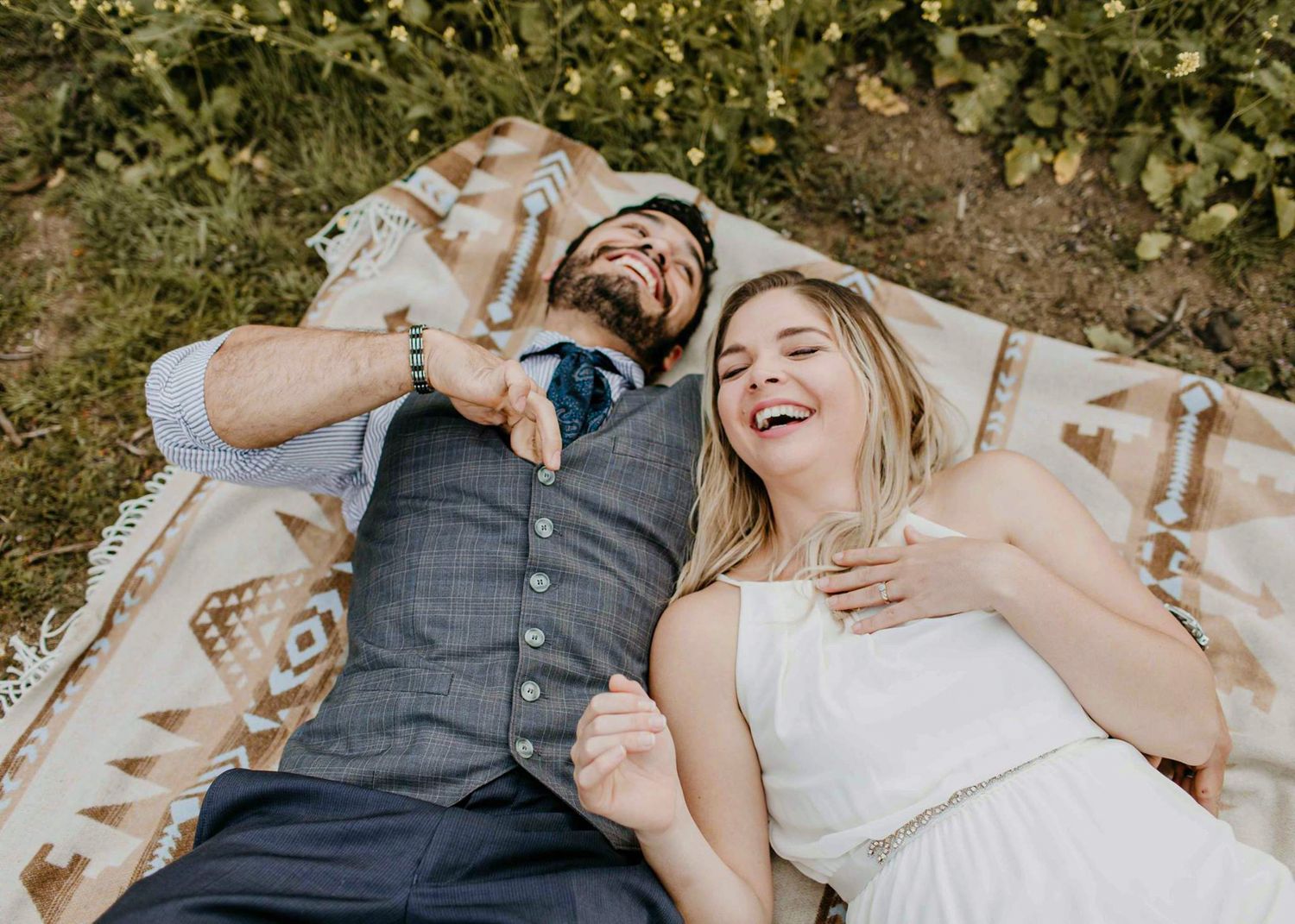 Happy couple laughing while lying on a patterned blanket outdoors in a sunny field.
