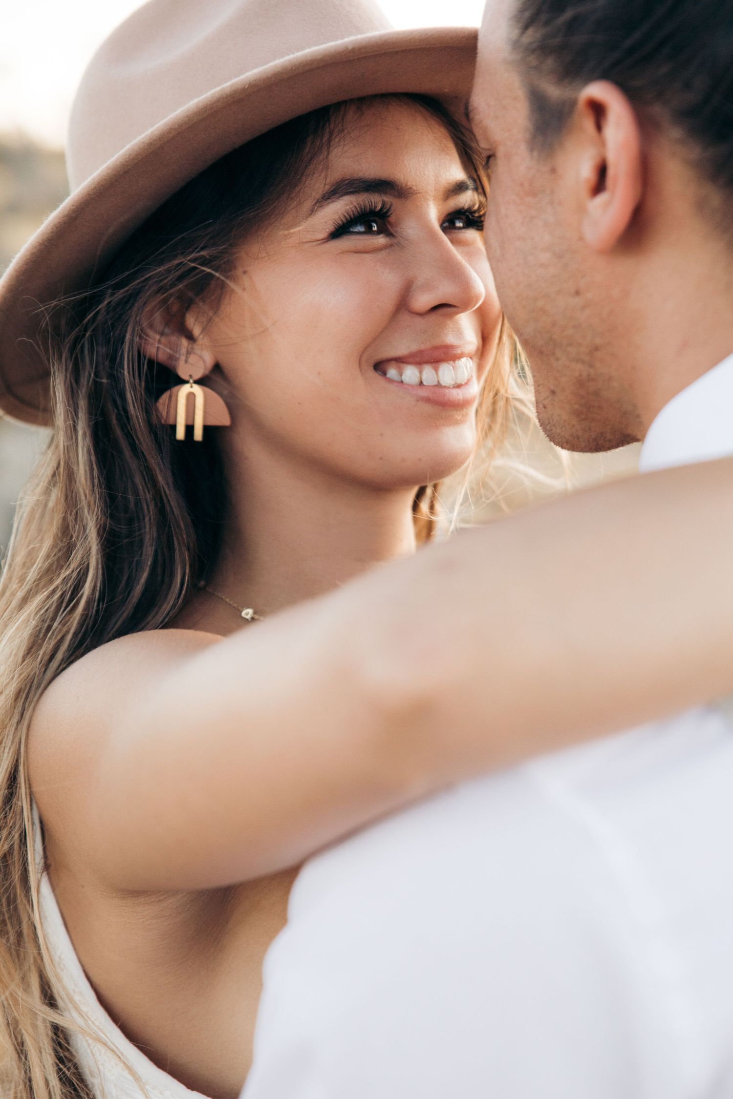 A smiling woman in a beige hat and geometric earrings gazes lovingly at her partner outdoors.