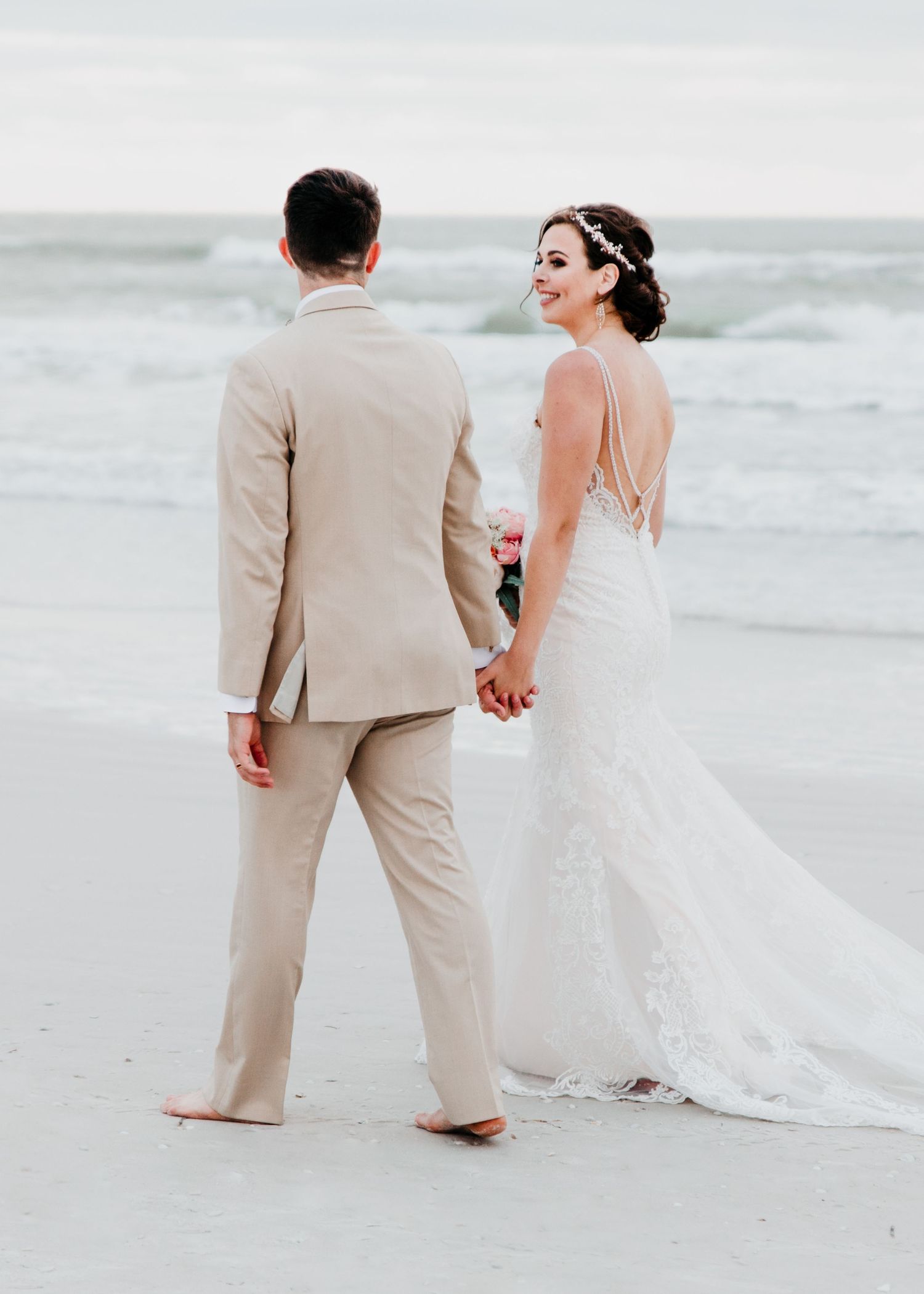 Bride and groom holding hands on sandy beach with ocean waves in background.