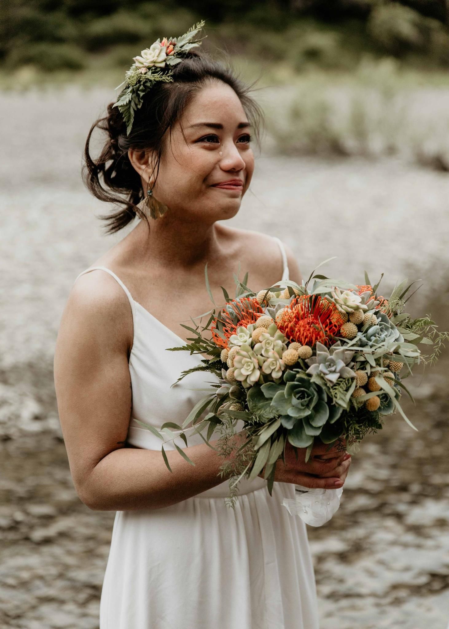 Tearful bride in white dress holding colorful bouquet with succulents and orange flowers, wearing floral crown.