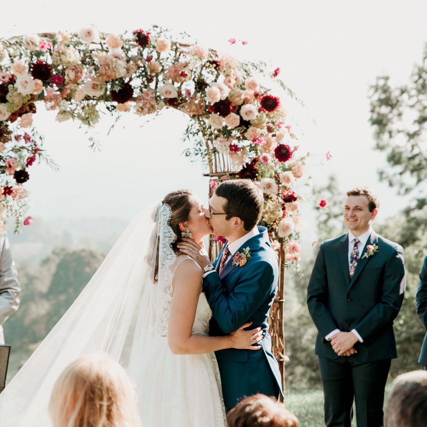Bride and groom sharing their first kiss under a stunning floral arch at an outdoor mountain wedding ceremony.