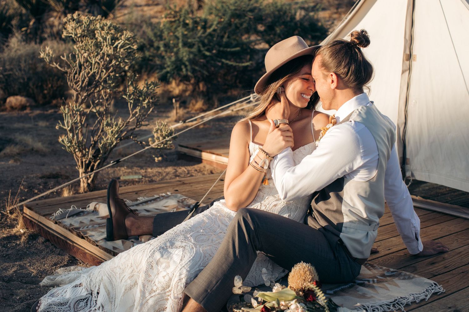 Romantic couple embracing outside a boho-style tent in a desert setting, bride in white lace dress and wide-brim hat.