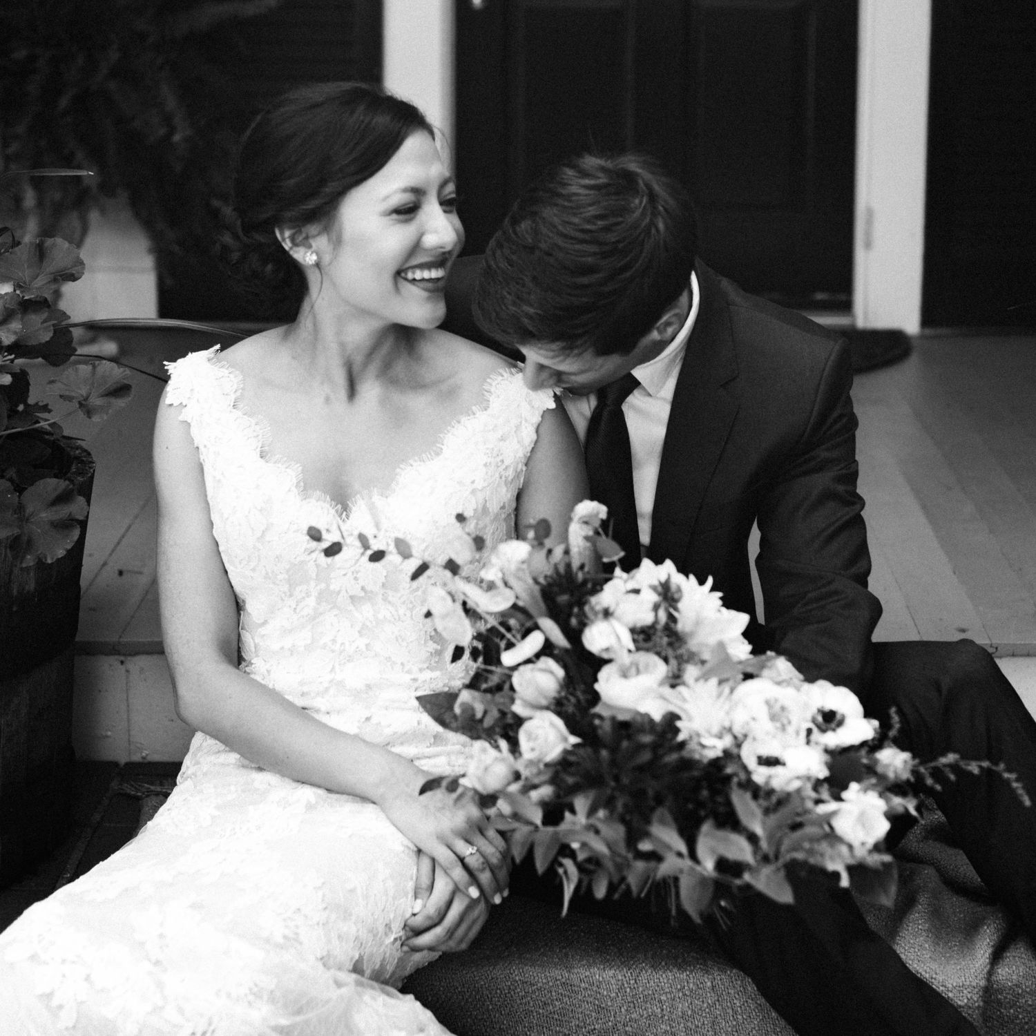 Black and white photo of laughing bride and groom sitting together with bouquet.