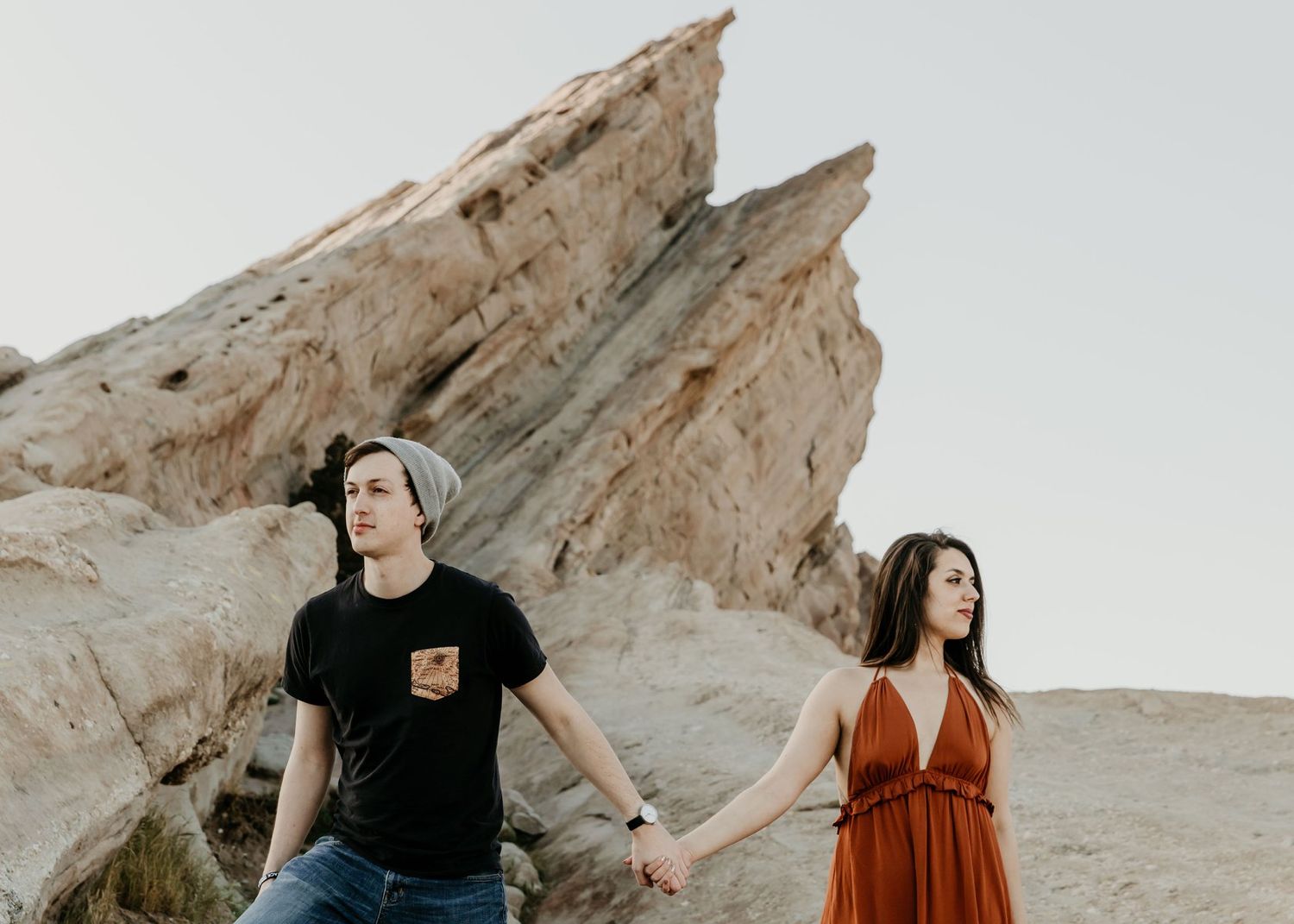 Couple holding hands at Vasquez Rocks, woman in rust dress and man in black tee with gray beanie.