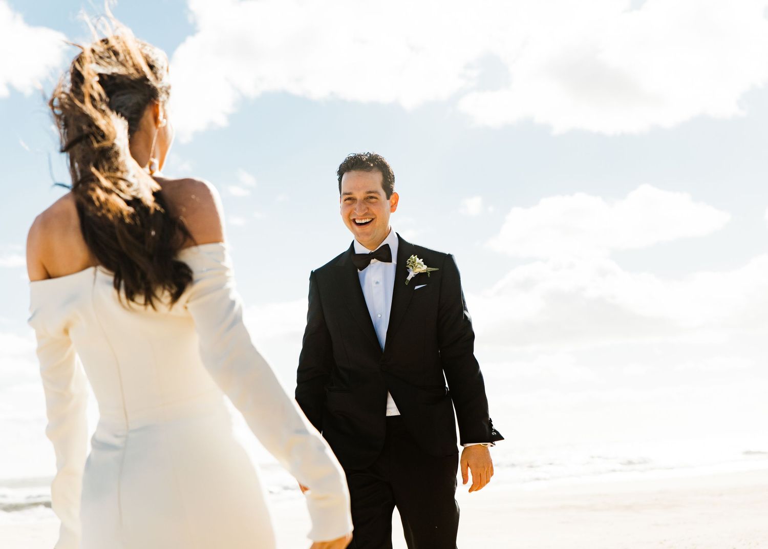 Smiling groom in black tuxedo seeing his bride for the first time on a sunny beach.