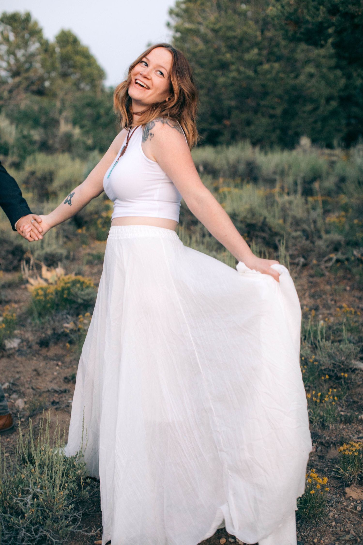 A joyful bride in a two-piece white tulle skirt twirls while holding hands in a natural outdoor setting.