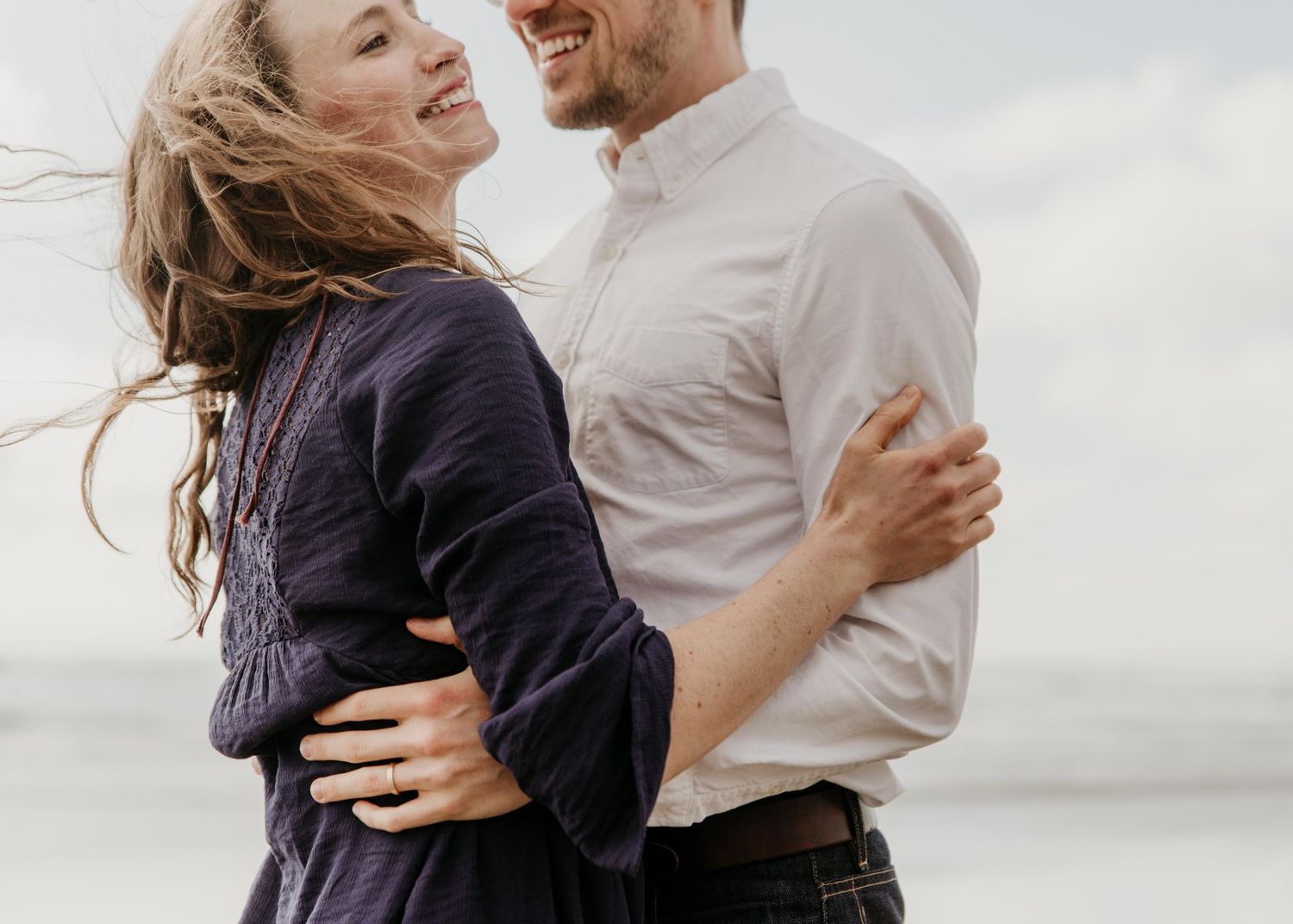 A happy couple embraces and laughs together on a windy beach with hair blowing in the breeze.