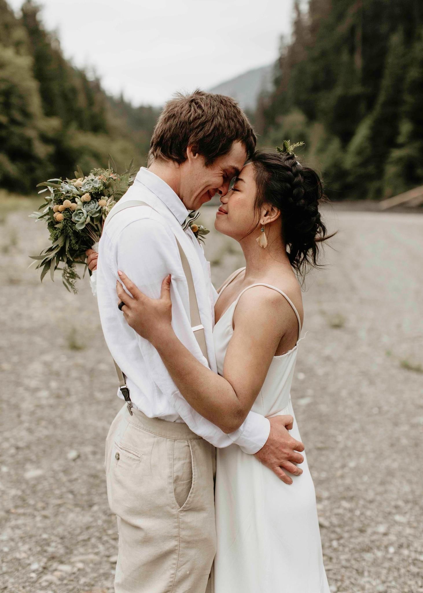 Bride in white slip dress and groom in suspenders sharing a kiss in a mountain valley.