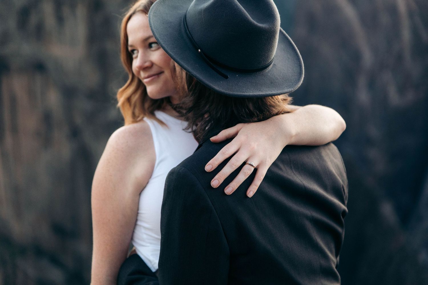 A smiling woman hugs a man in a black hat and jacket against a rocky outdoor backdrop.
