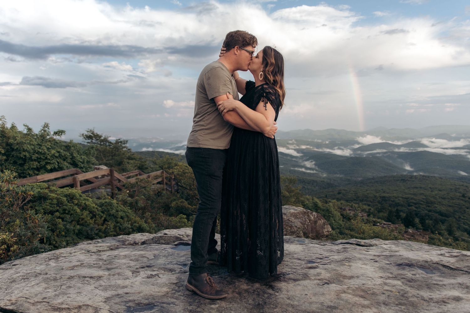 Couple kissing on a rocky overlook with a rainbow and misty mountain valley behind them.