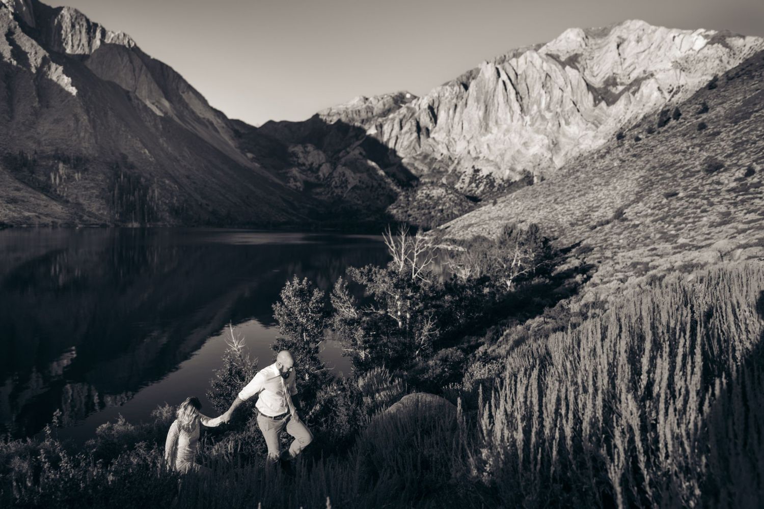 Black and white photo of a couple holding hands by a reflective mountain lake surrounded by dramatic peaks.