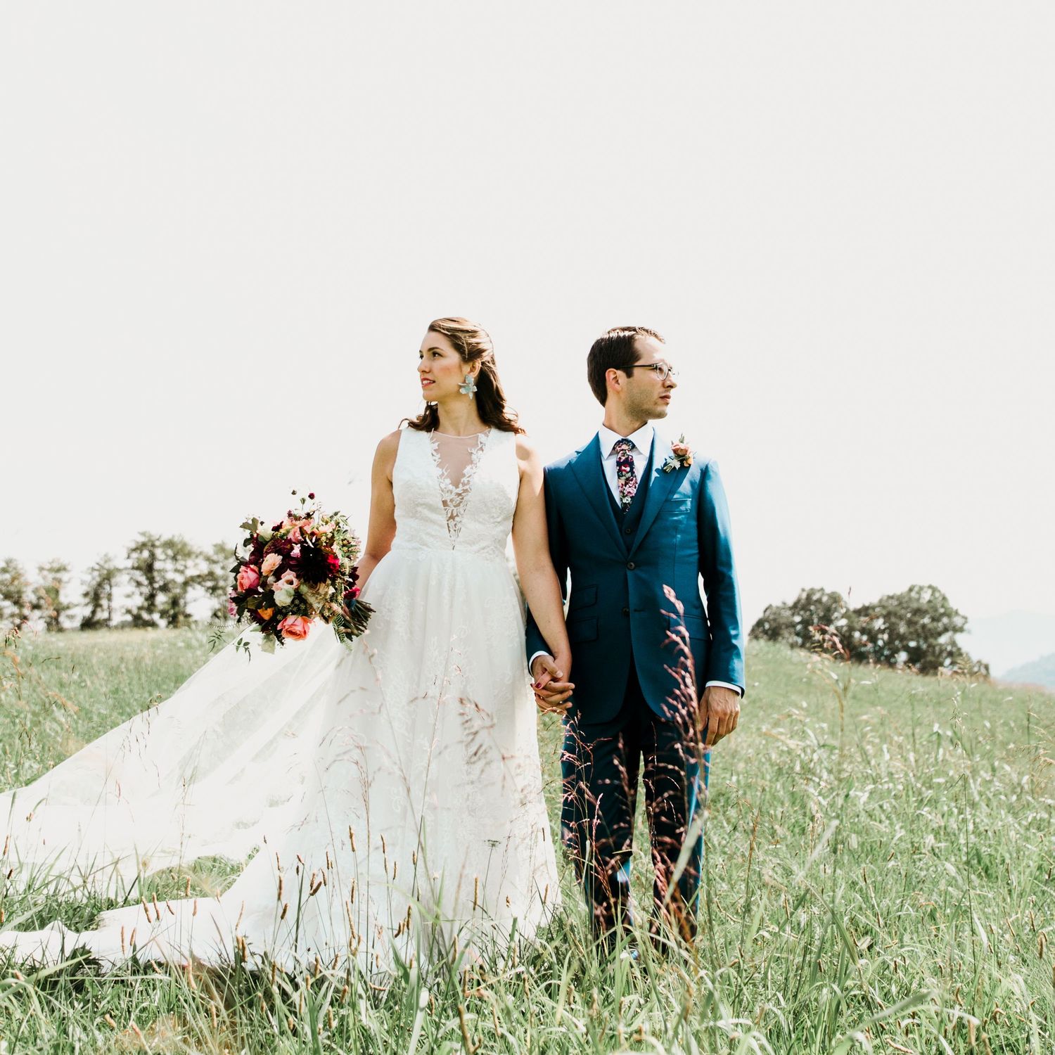 Bride and groom stand hand in hand in a sunlit meadow, bride holding a vibrant red and pink bouquet.